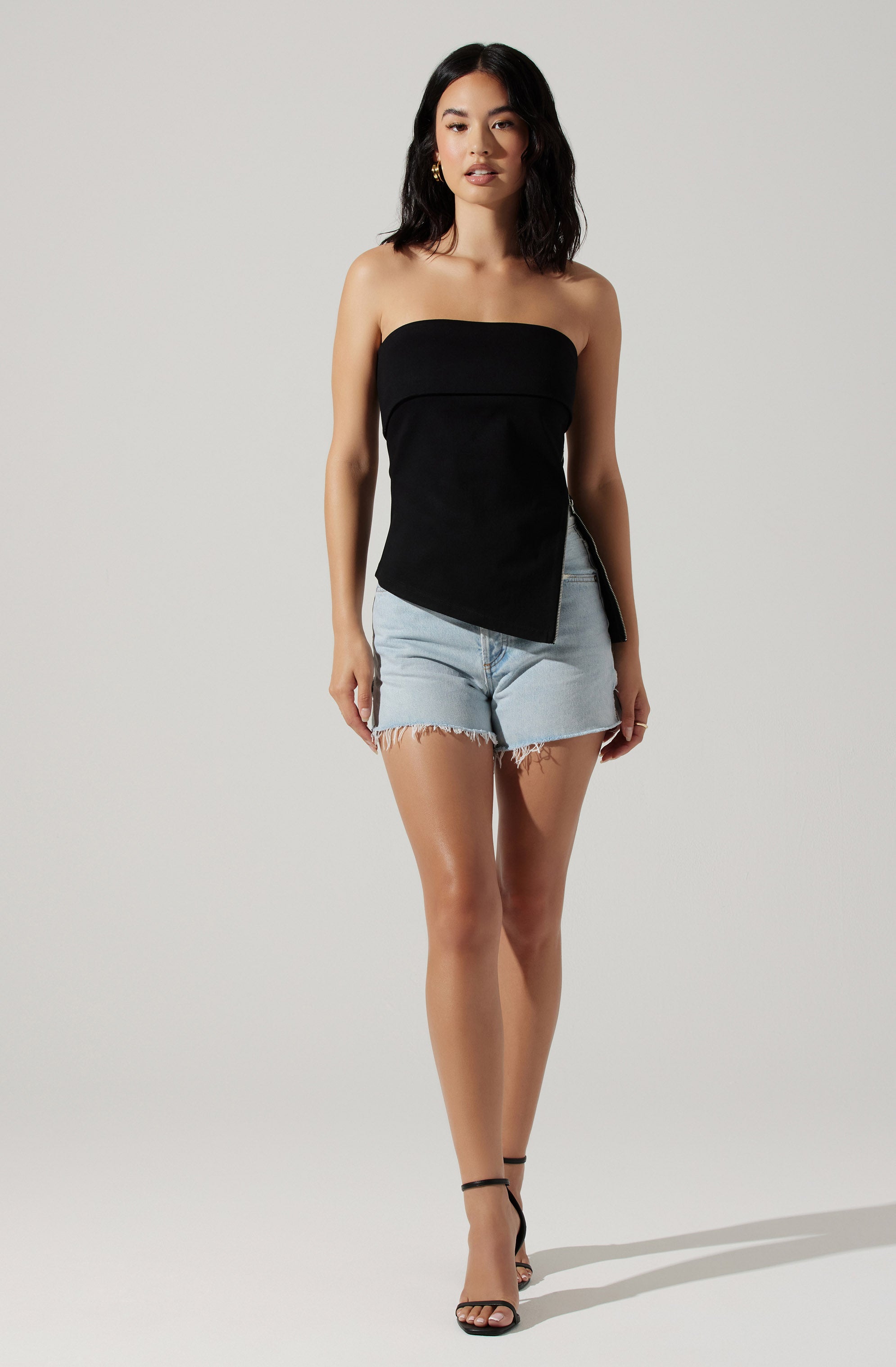 A woman is wearing a strapless black top with an asymmetrical hemline that is longer on one side, paired with light-wash, high-waisted denim shorts with frayed hems. She is also wearing black, open-toe high-heeled sandals with ankle straps.