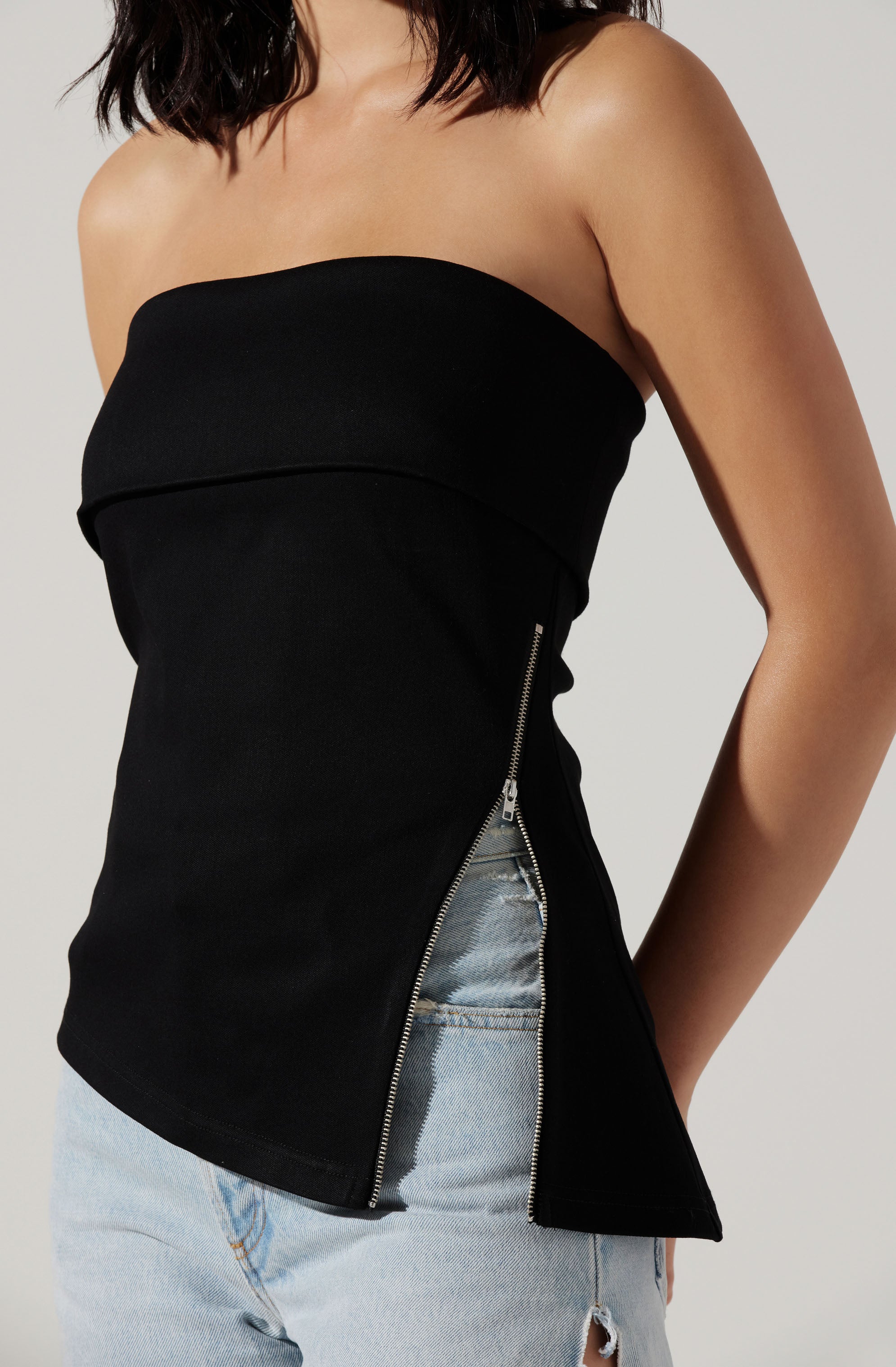 A woman is wearing a black strapless top with an asymmetrical zipper design on the side. The top appears fitted and ends at the hips. She is also wearing light-wash, distressed jeans with a ripped knee.