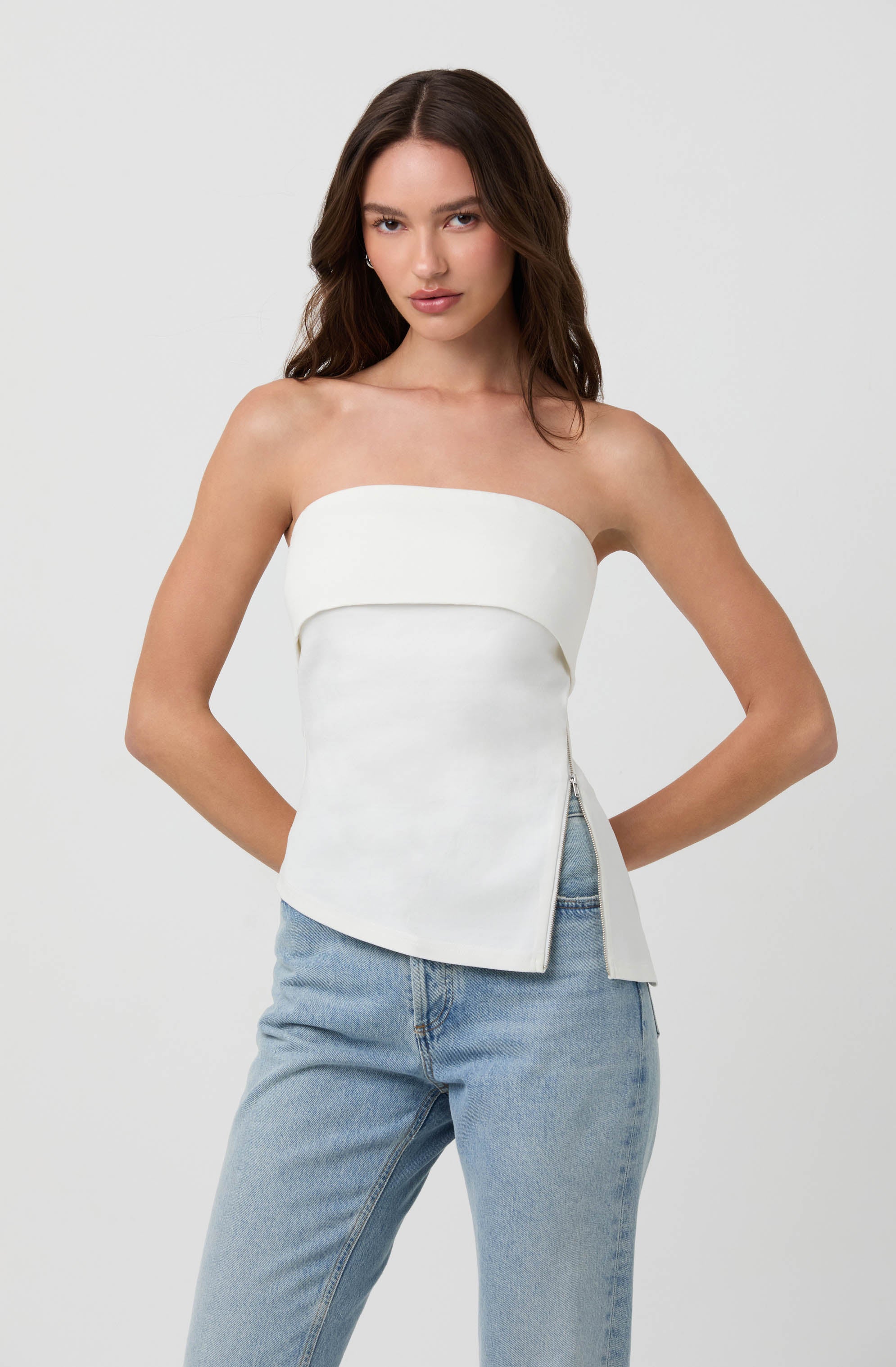 A woman is wearing a strapless white top with a horizontal band across the chest and a fitted, slightly asymmetrical hem. She pairs it with light blue, high-waisted jeans.