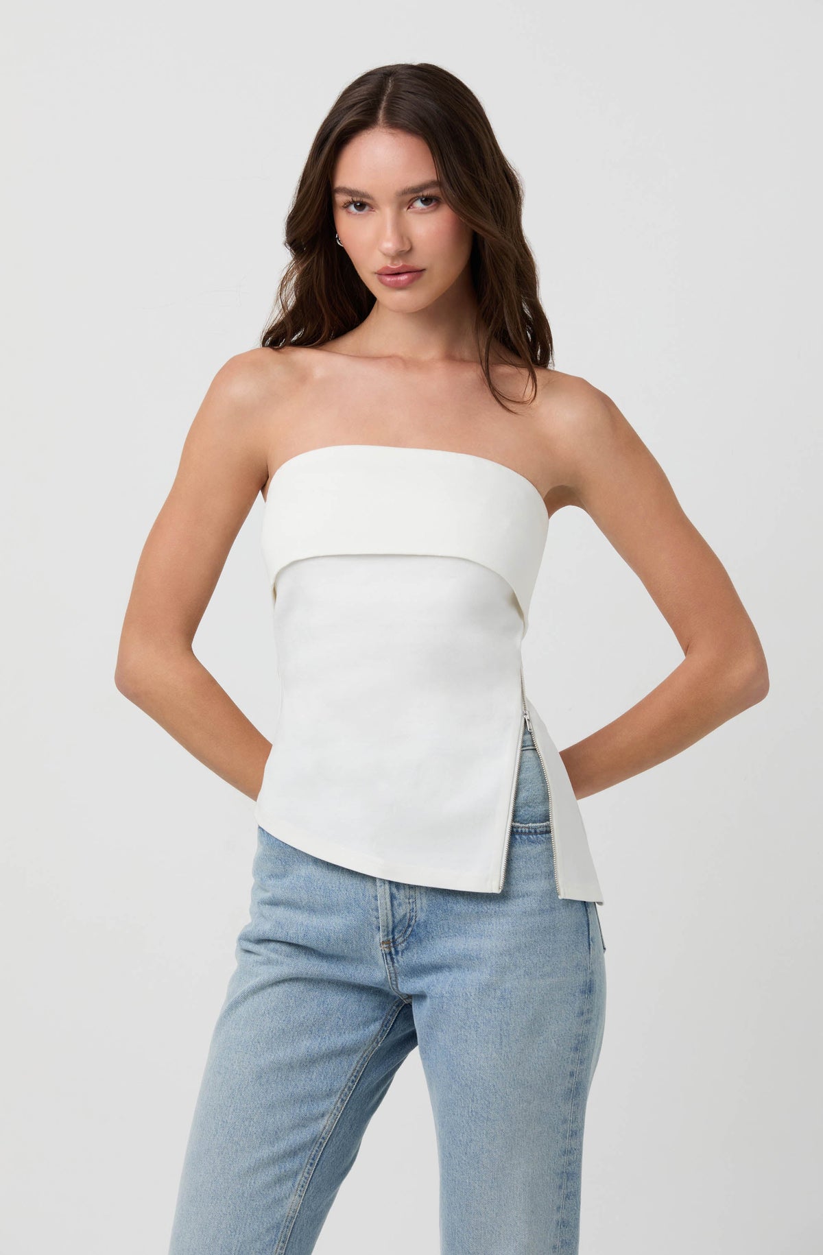 A woman is wearing a strapless white top with a horizontal band across the chest and a fitted, slightly asymmetrical hem. She pairs it with light blue, high-waisted jeans.