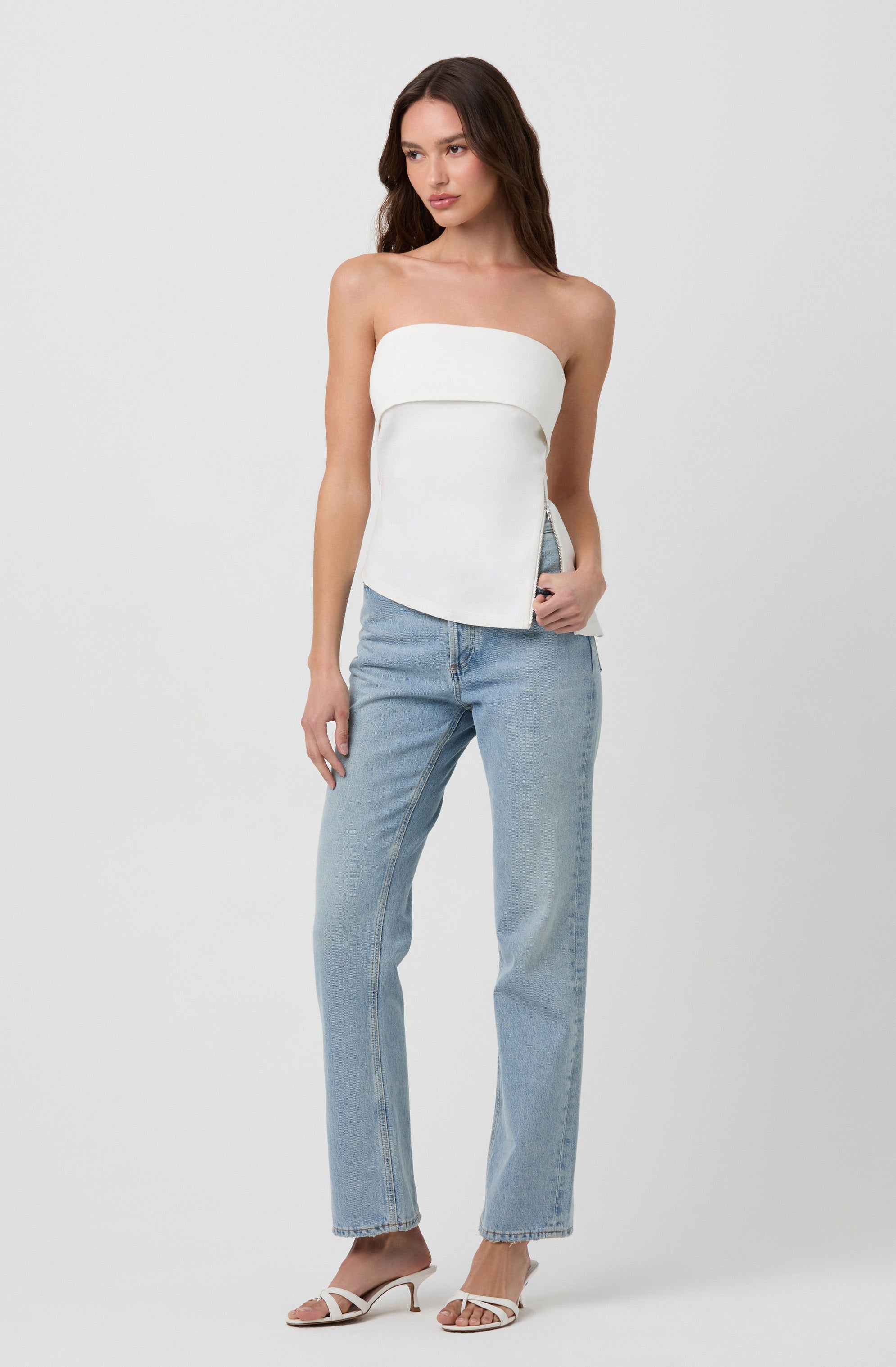 A woman is wearing a strapless white top with a layered design and light blue high-waisted jeans. She has on white heeled sandals with thin straps.