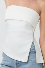 A woman is wearing a strapless white top with an asymmetrical design. The top has a curved, layered fabric detail across the top edge, and a silver zipper runs vertically along the side. She is also wearing light blue denim jeans.