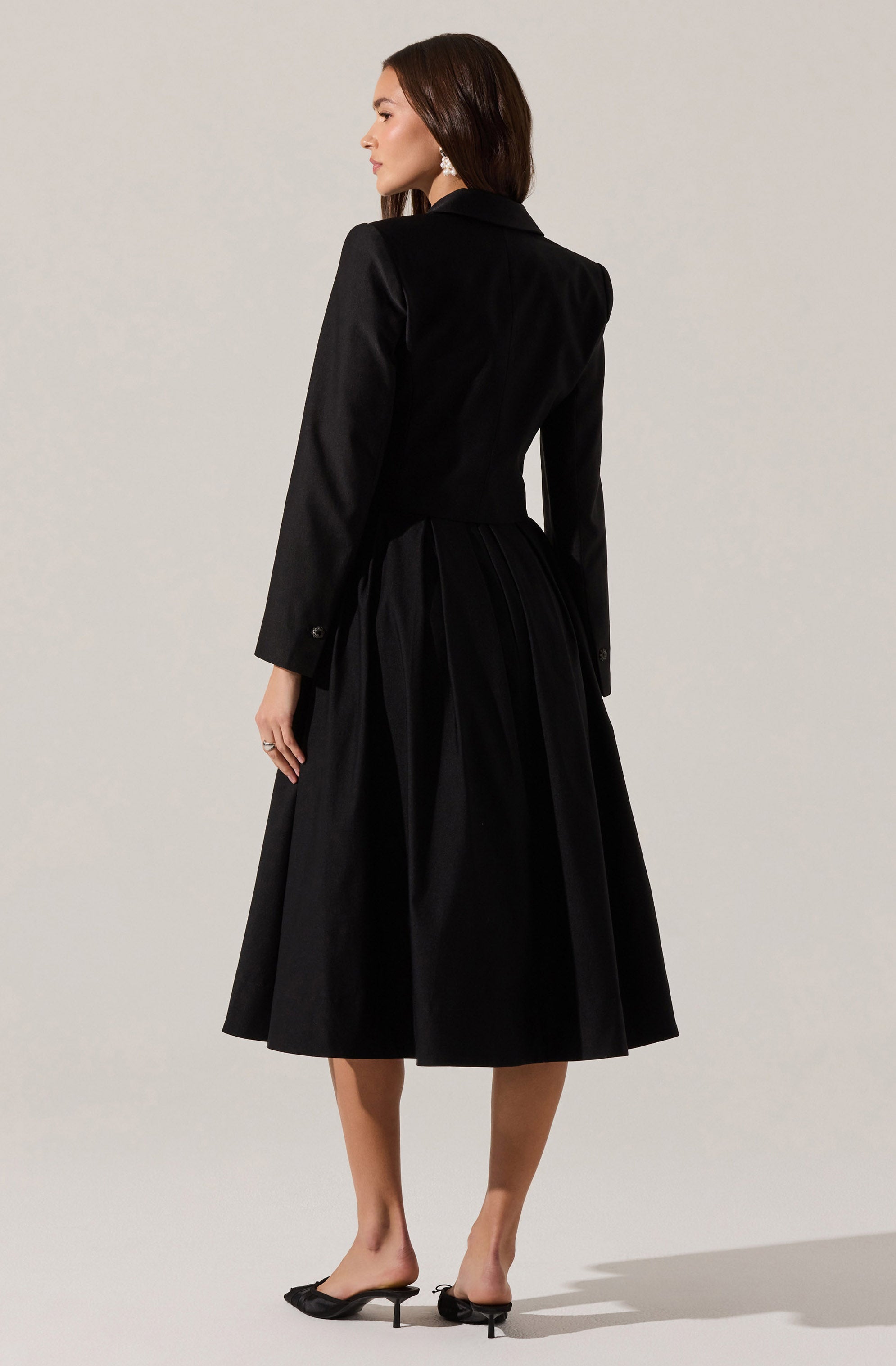 A woman is wearing a black blazer with long sleeves and a fitted waist, paired with a matching black midi-length skirt that has pleats and a full silhouette. She is also wearing black open-toe mules with small heels.