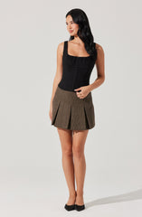 A woman is wearing a black sleeveless top with a gathered neckline and front button details. She pairs it with a high-waisted, brown pleated skirt with a subtle checkered pattern. She completes the outfit with black pointed-toe flats featuring a bow embellishment.