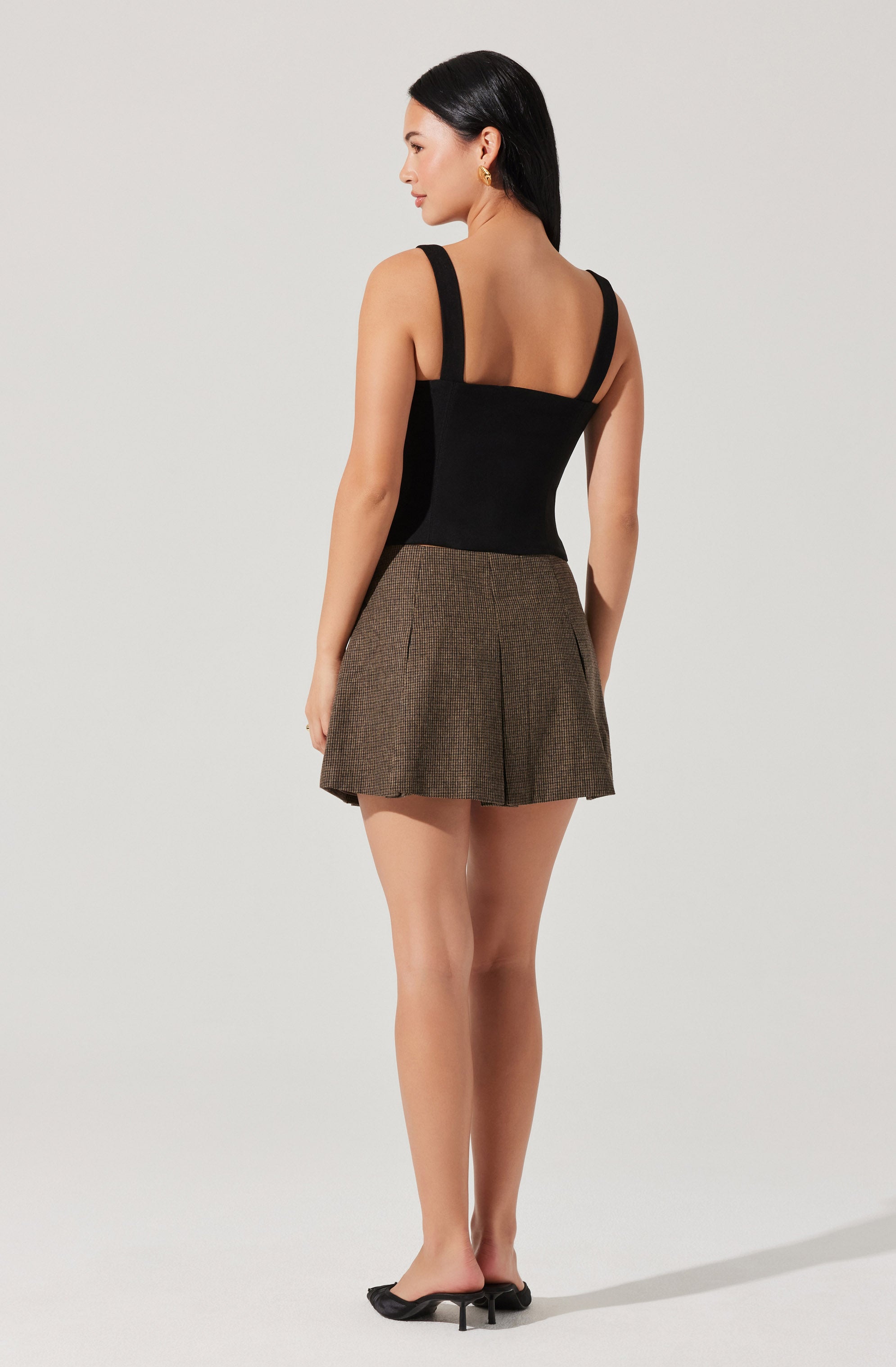 A woman is wearing a black sleeveless top with wide straps and a fitted bodice, paired with high-waisted, brown plaid shorts featuring pleats. She is also wearing black high-heeled shoes.