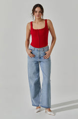 A woman wearing a fitted red sleeveless top with a square neckline and button detail down the front, paired with high-waisted light blue wide-leg jeans and white heels.