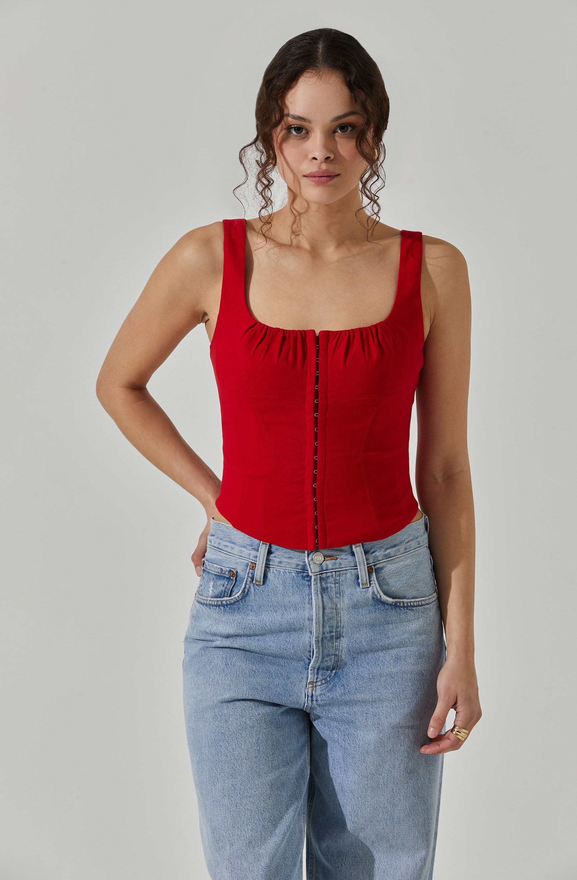 A woman is wearing a sleeveless red top with a square neckline and gathered fabric detail at the front, paired with light blue high-waisted jeans.