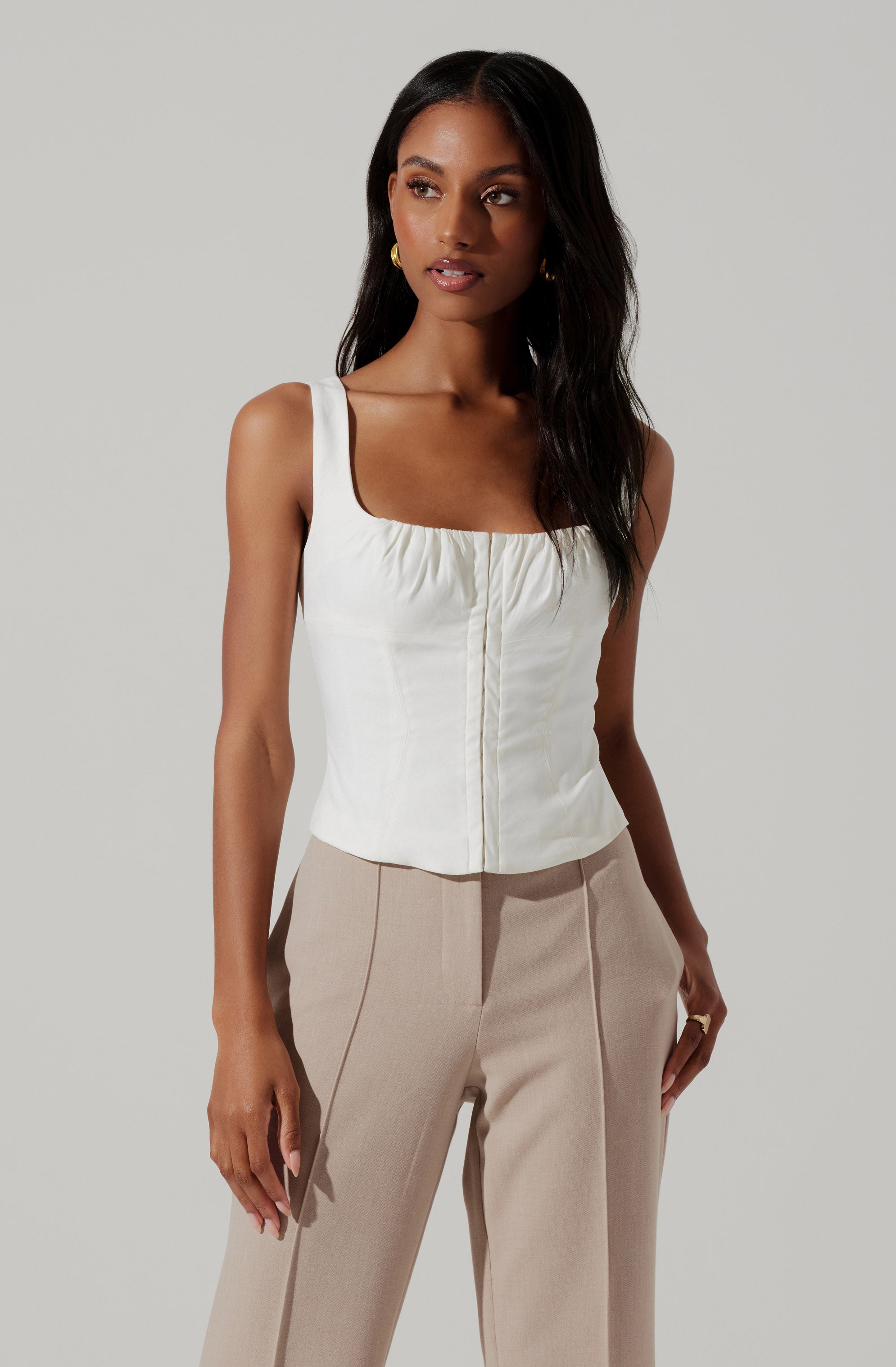 A woman is wearing a sleeveless, cream-colored top with wide straps and a square neckline, featuring a gathered detail along the neckline and a vertical seam down the center. She pairs it with high-waisted, light beige trousers that have a tailored fit with visible vertical seam lines on the front.