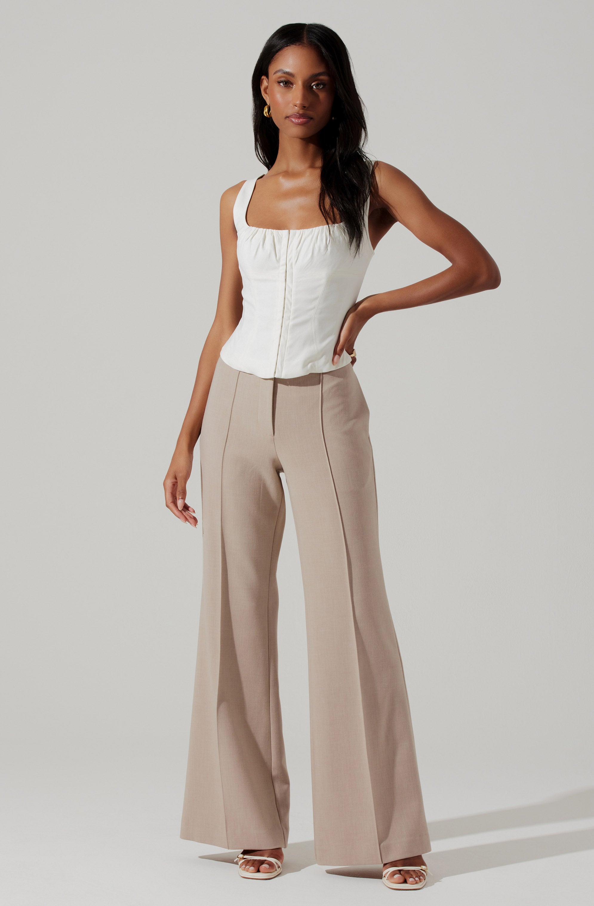 The woman is wearing a sleeveless, white top with a square neckline and a centered front zipper. She is also wearing high-waisted, wide-leg beige trousers with vertical seams running down the front. She accessorizes with gold hoop earrings and is wearing open-toe heels.