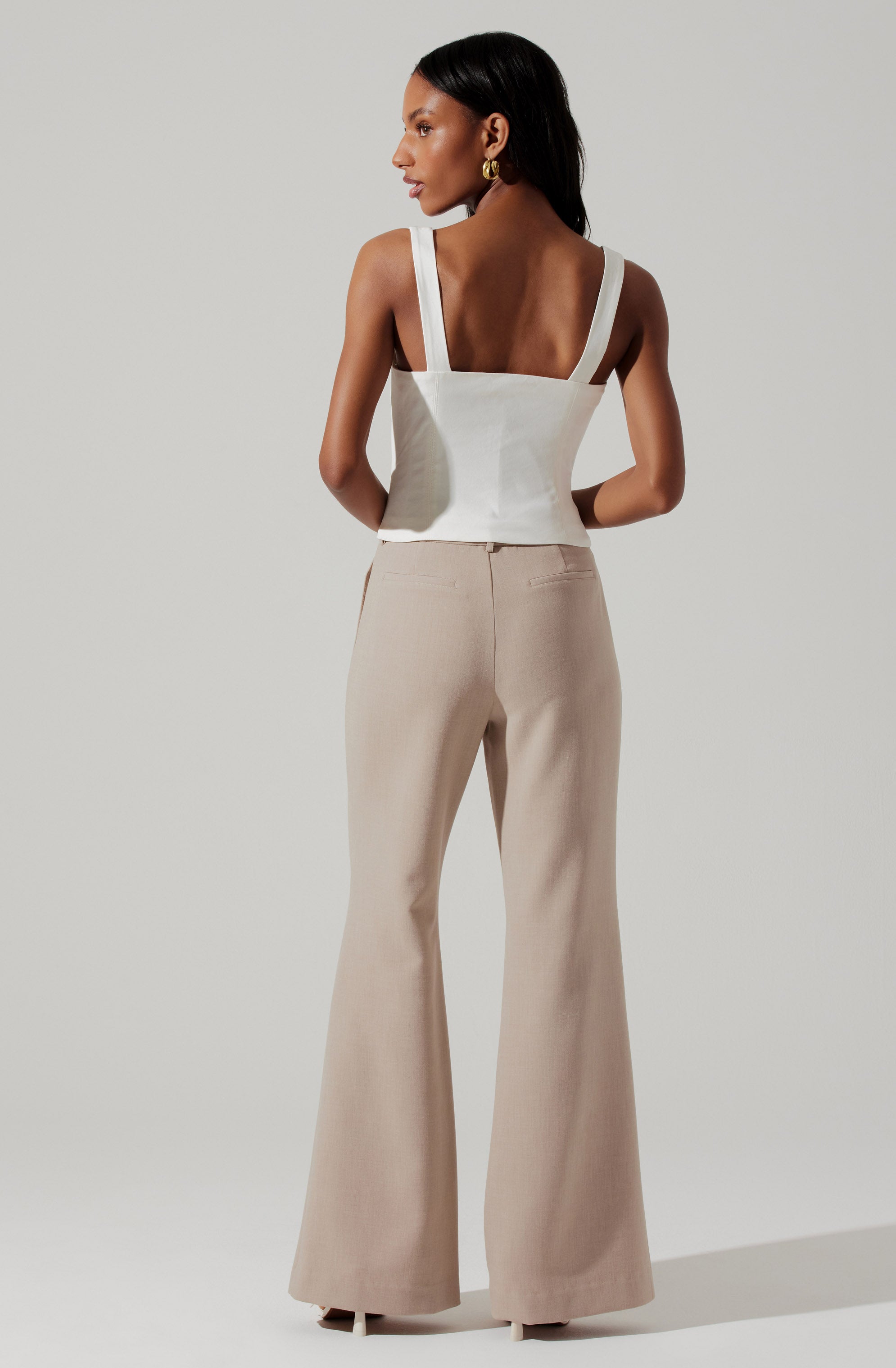 A woman is wearing a white sleeveless top with wide shoulder straps and a square neckline. She pairs it with high-waisted, beige, wide-leg pants that flare out at the bottom.