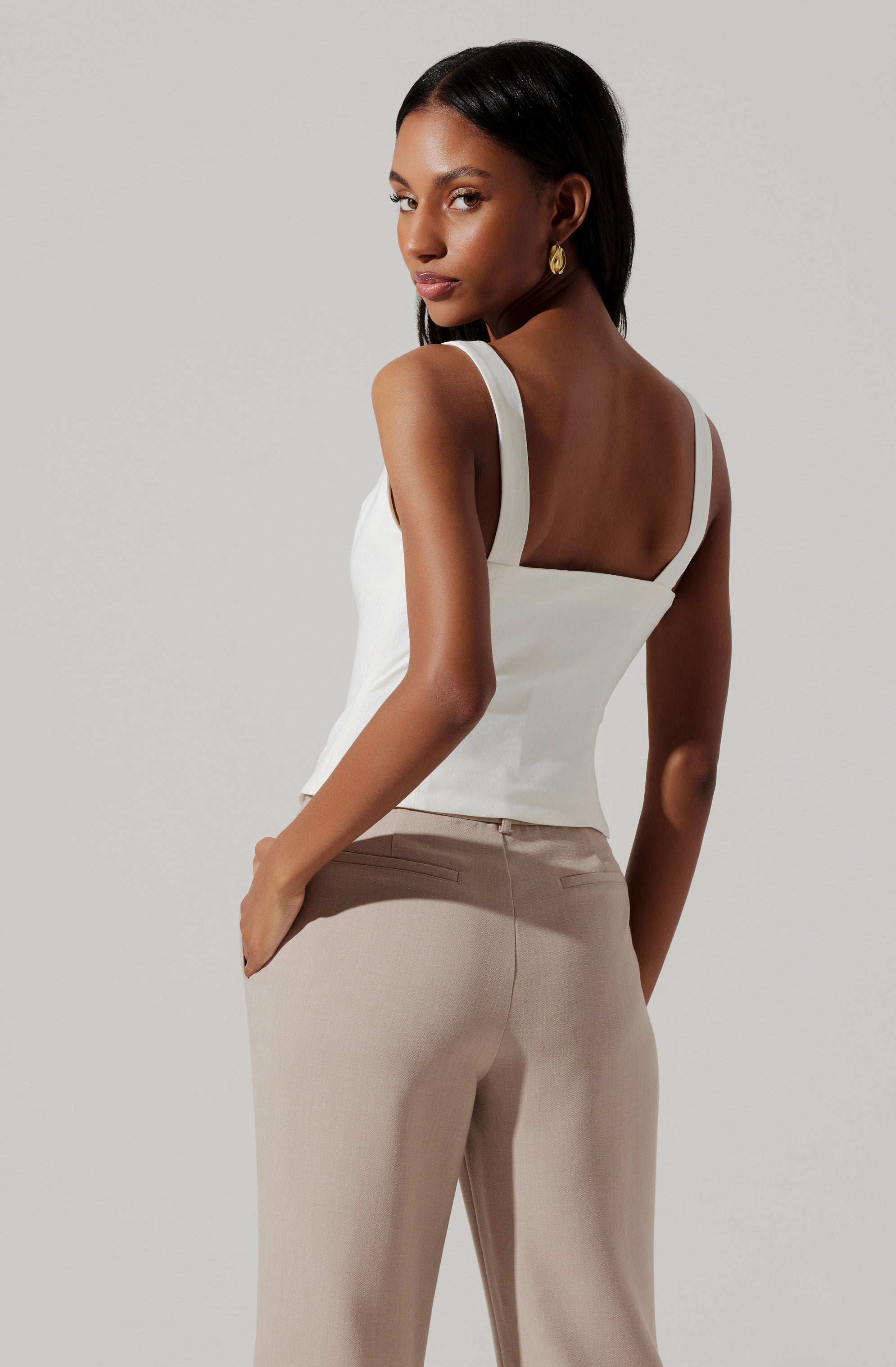 A woman is wearing a white sleeveless top with wide straps and a fitted bodice. She is also wearing beige, high-waisted, tailored pants with a smooth, flat front.