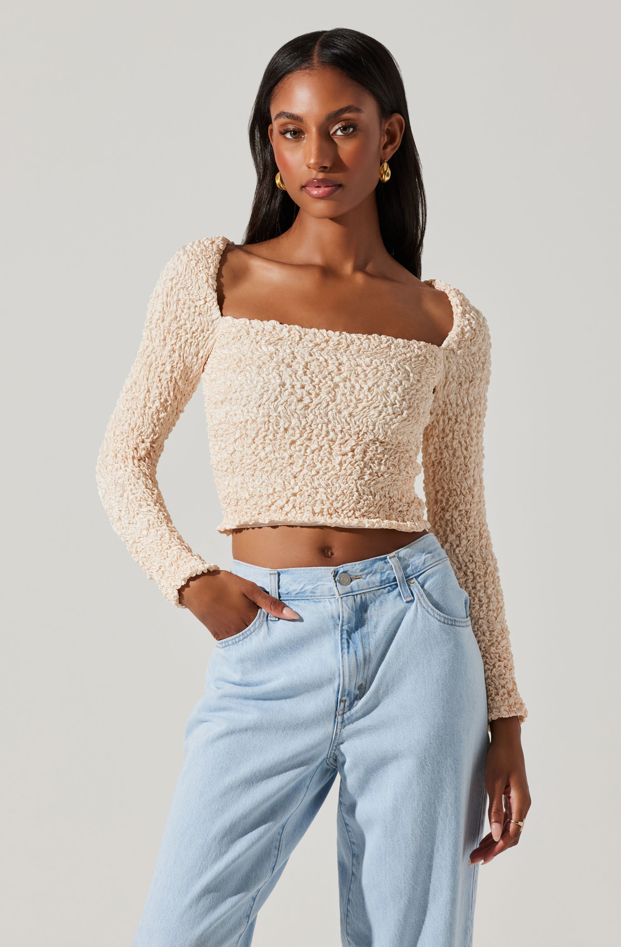A woman is wearing a beige, textured, long-sleeve, crop top with a square neckline paired with light-wash high-waisted jeans. She accessorized with gold hoop earrings.