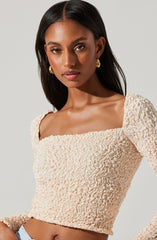 A woman is wearing a long-sleeved, textured cream-colored crop top with a square neckline. She has paired it with gold hoop earrings.