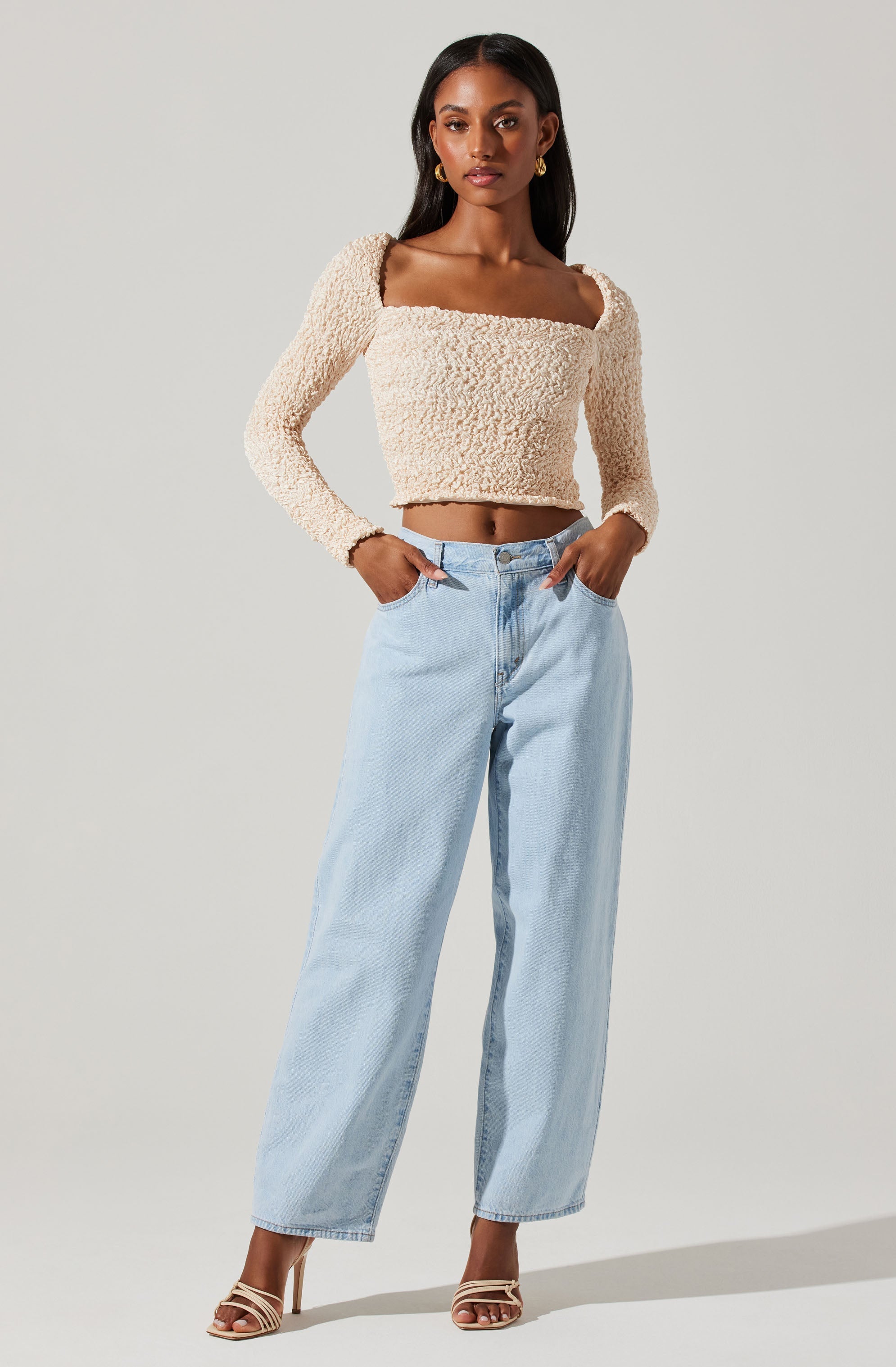 A woman is wearing a beige, textured, long-sleeve crop top with a square neckline, paired with high-waisted, light wash wide-leg jeans. She is also wearing beige strappy high-heeled sandals and gold hoop earrings.