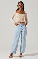 A woman is wearing a beige, textured, long-sleeve crop top with a square neckline, paired with high-waisted, light wash wide-leg jeans. She is also wearing beige strappy high-heeled sandals and gold hoop earrings.
