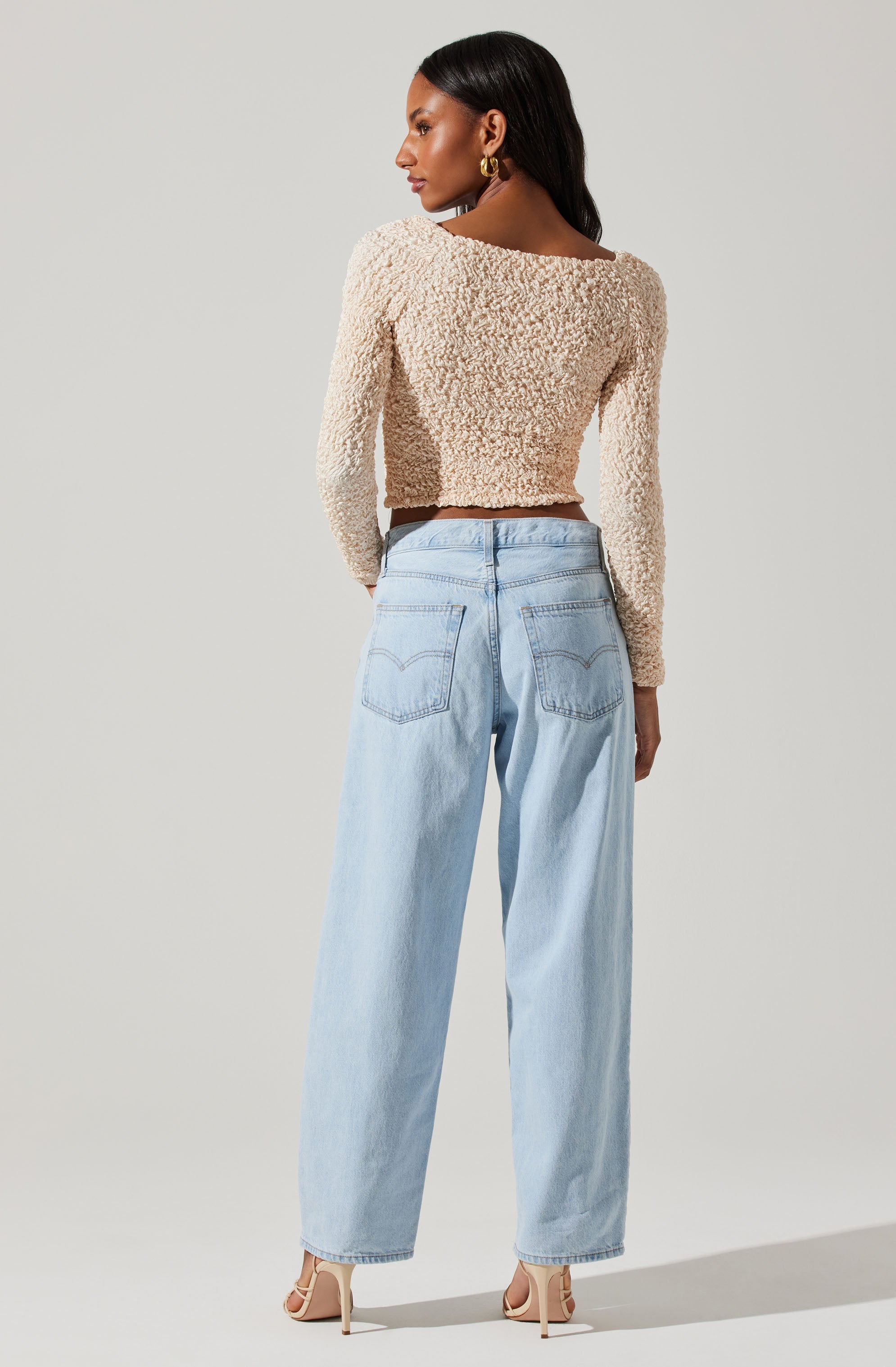 A woman is wearing a cream-colored, textured, long-sleeve cropped sweater paired with high-waisted, light-wash wide-leg jeans. She also has on gold hoop earrings and nude high-heeled shoes.