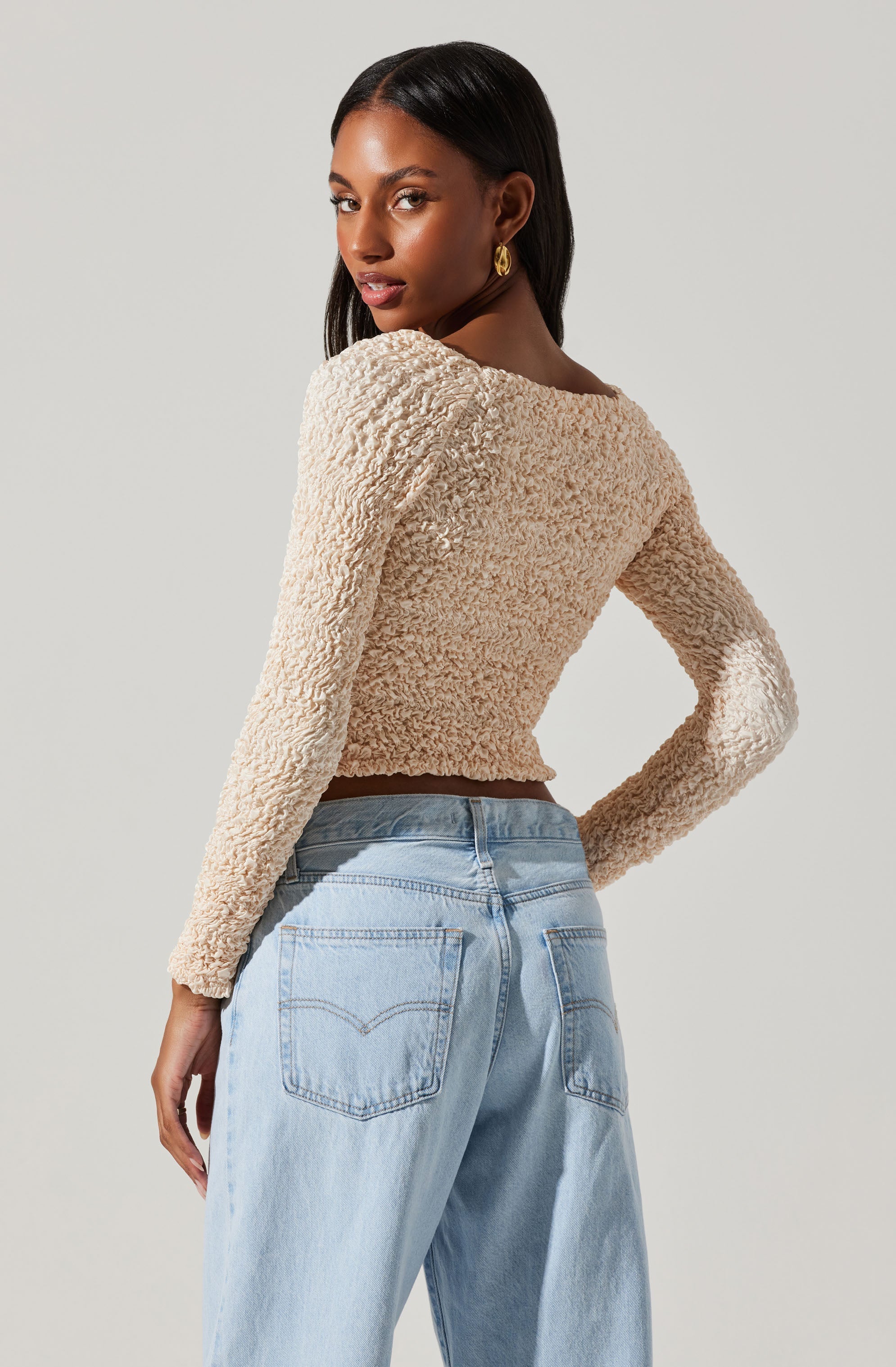 A woman is wearing a long-sleeved, textured, beige cropped sweater and light blue high-waisted jeans.