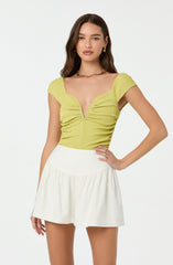 A young woman is wearing a lime green, textured, short-sleeved top with a deep V neckline and gathered detailing at the front. She pairs it with a high-waisted, white, slightly A-line skirt that features a dropped waistband and gentle gathers. She accessorizes with gold earrings and a gold chain necklace.