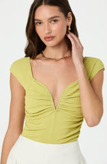 A woman is wearing a yellow, textured, short-sleeved top with a deep V neckline. The top has a ruched, gathered design at the front. She is also wearing a gold chain necklace and matching gold earrings.