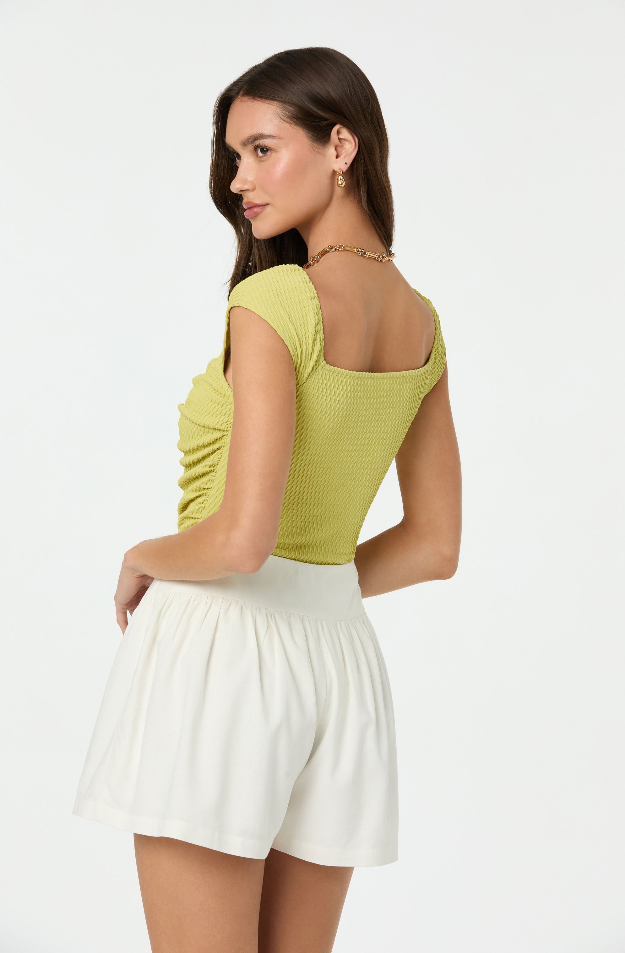 A woman is wearing a short-sleeved, light yellow, textured top with a square neckline, paired with high-waisted, loose-fitting white shorts. She has long, dark brown hair and is accessorized with gold earrings and a gold chain necklace.