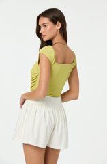 A woman is wearing a short-sleeved, light yellow, textured top with a square neckline, paired with high-waisted, loose-fitting white shorts. She has long, dark brown hair and is accessorized with gold earrings and a gold chain necklace.