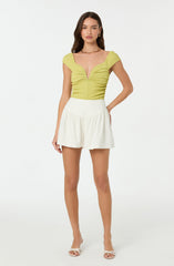 A woman is wearing a bright yellow, textured, short-sleeved top with a deep V neckline and ruched detailing in the center. She pairs it with high-waisted, white, flowy shorts and white open-toe heels. She accessorizes with gold earrings and a gold chain necklace.
