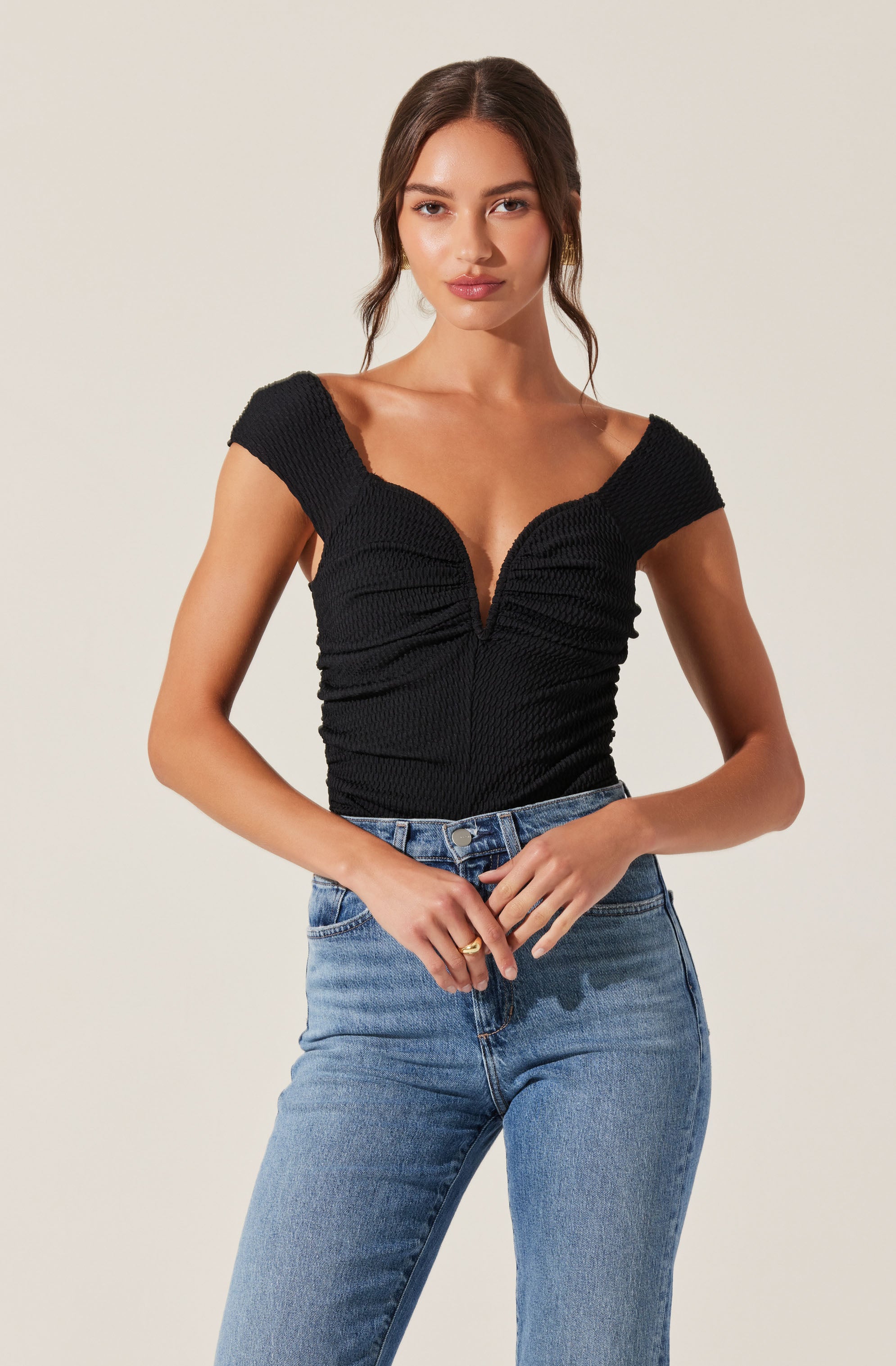 A woman is wearing a fitted black top with a deep V-neckline and short, wide sleeves. The top features a textured, ribbed fabric with ruched detailing along the front and sides. She pairs it with high-waisted, light blue denim jeans.