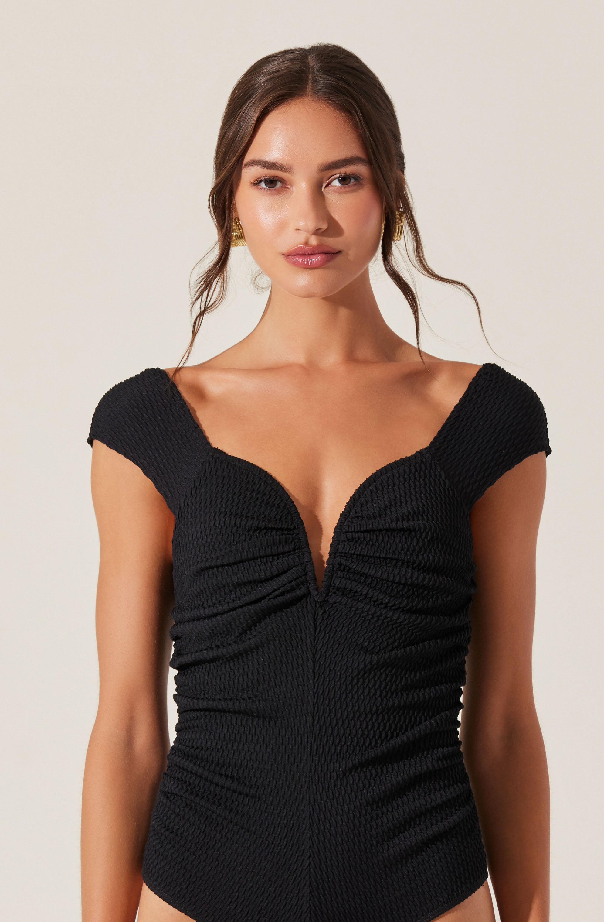 A woman is wearing a sleek black sleeveless top with a deep V neckline. The fabric has a textured, ruched appearance that creates a fitted silhouette. The top features wide shoulder straps and gathers at the front that accentuate the bust.