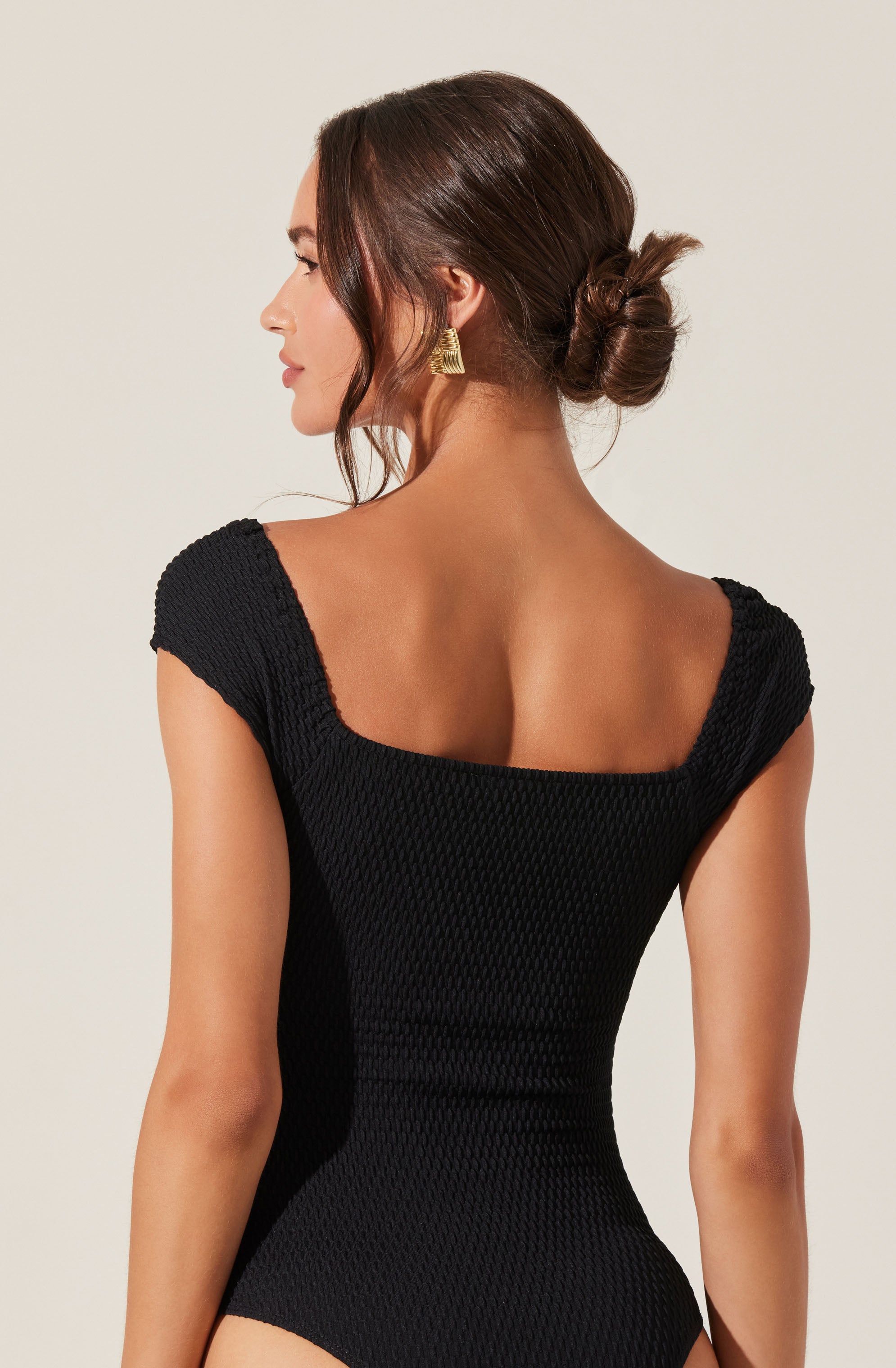 A woman is wearing a black textured dress with short, gathered sleeves and a square neckline. She has her hair styled in a low, twisted bun and is accessorized with large, gold, triangular earrings.