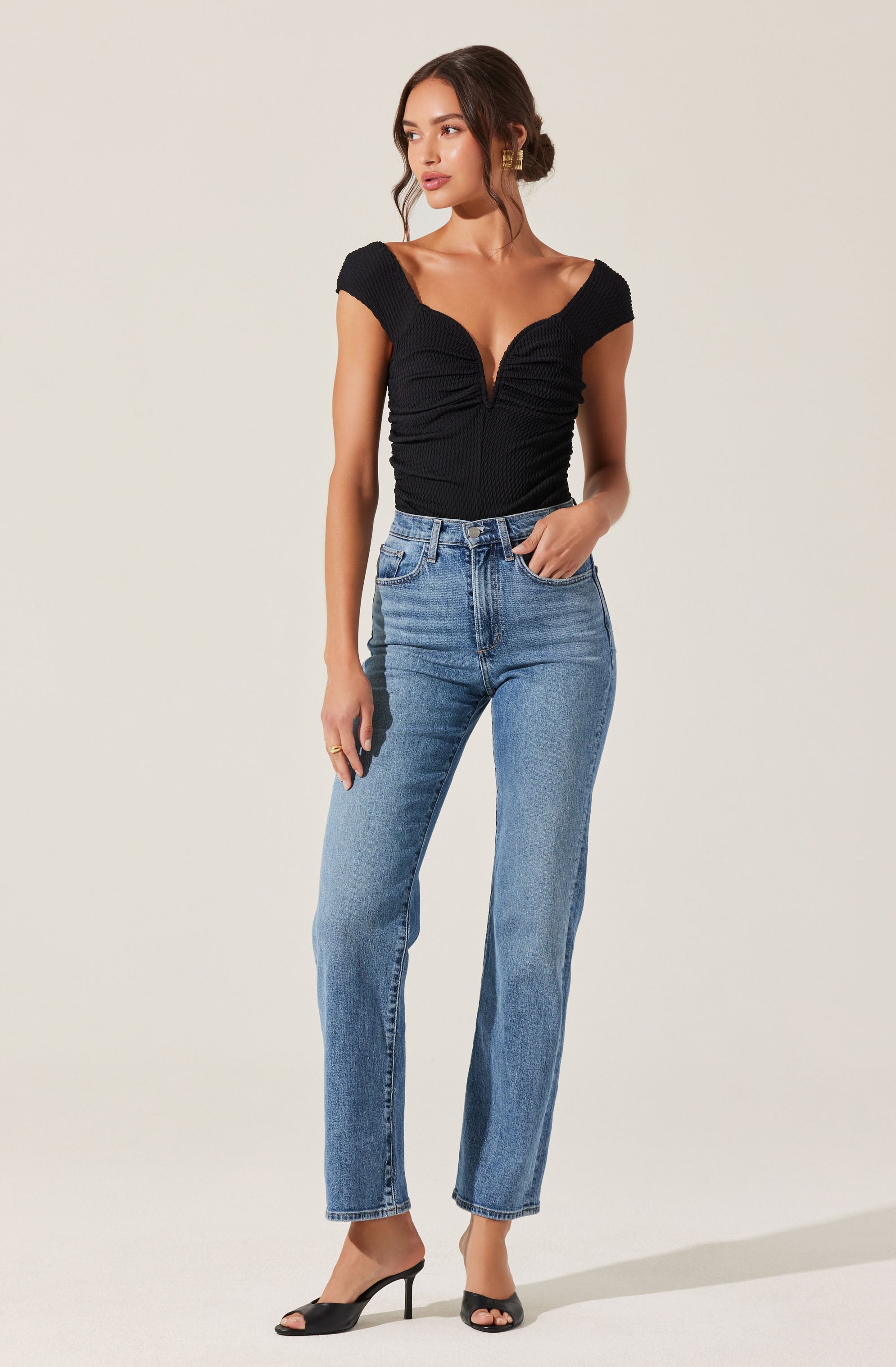 A woman is wearing a black, short-sleeved top with a deep V-neck and ruched detailing. She pairs it with high-waisted, light-wash blue jeans that have a straight-leg fit. She completes her outfit with black open-toe high-heeled shoes.