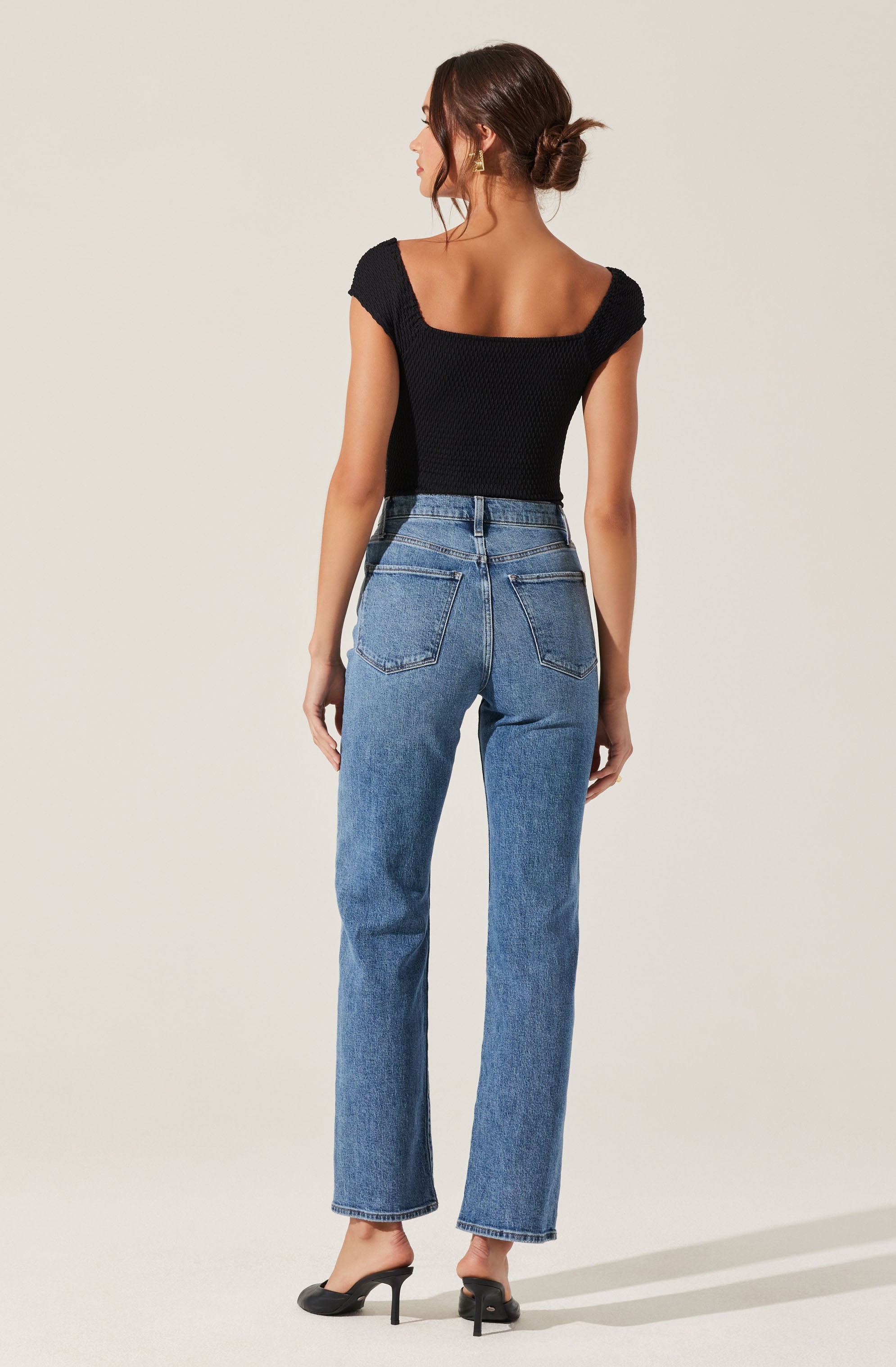 A woman is wearing a fitted black short-sleeve top with a textured knit pattern, featuring a wide, rounded neckline that extends to the shoulders. She pairs it with high-waisted, straight-leg blue jeans that have a classic five-pocket design, including two back pockets. She completes her outfit with black high-heeled sandals.