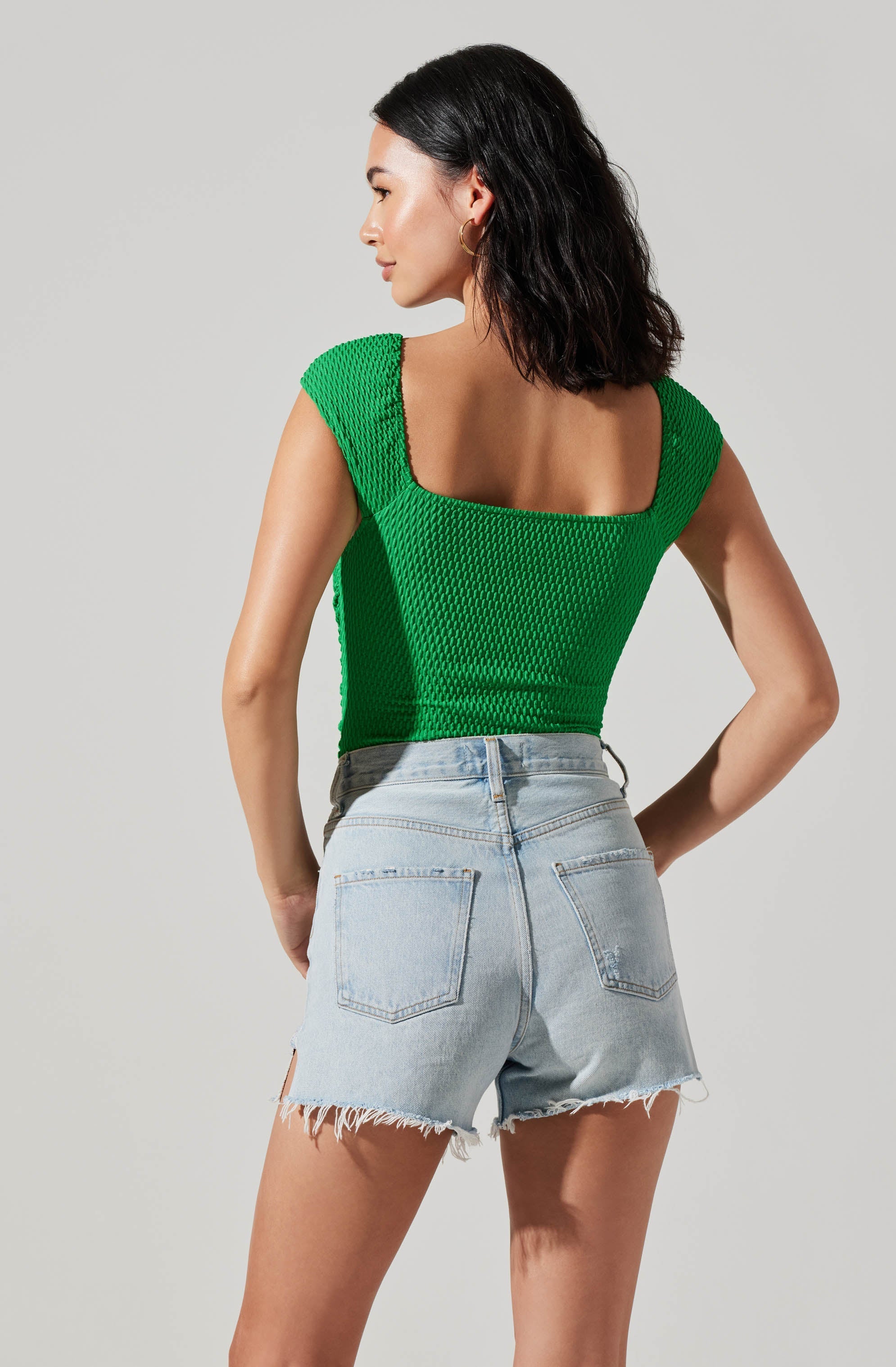 A woman wearing a bright green, textured sleeveless top with wide shoulder straps and a squared neckline, paired with high-waisted, light-wash denim shorts with frayed hems and back pockets.