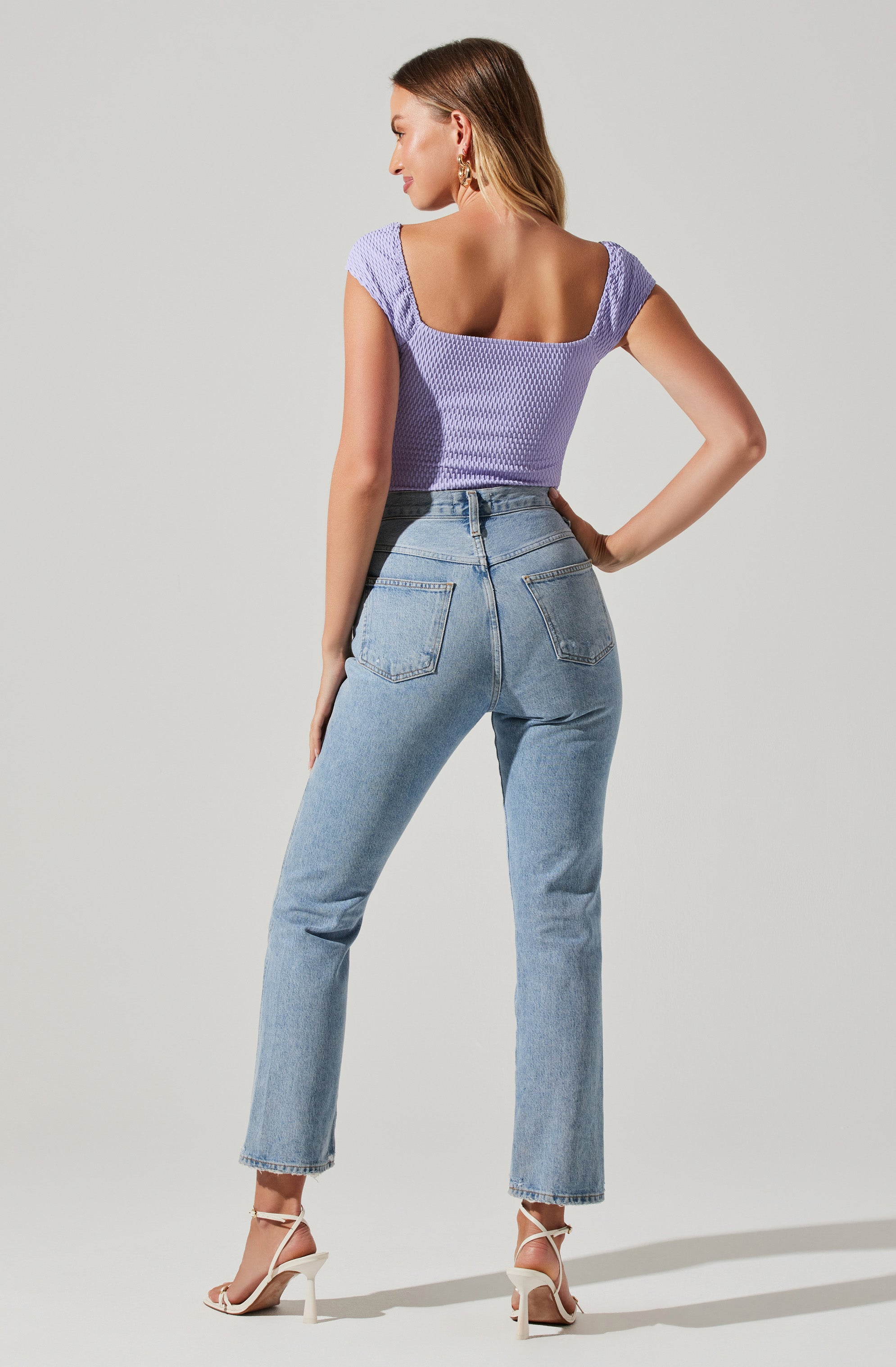 A woman is wearing a lavender short-sleeved, textured top with a scoop neckline, paired with high-waisted light-wash denim jeans that have a straight-leg fit and back pockets. She is also wearing beige strappy high-heeled sandals.