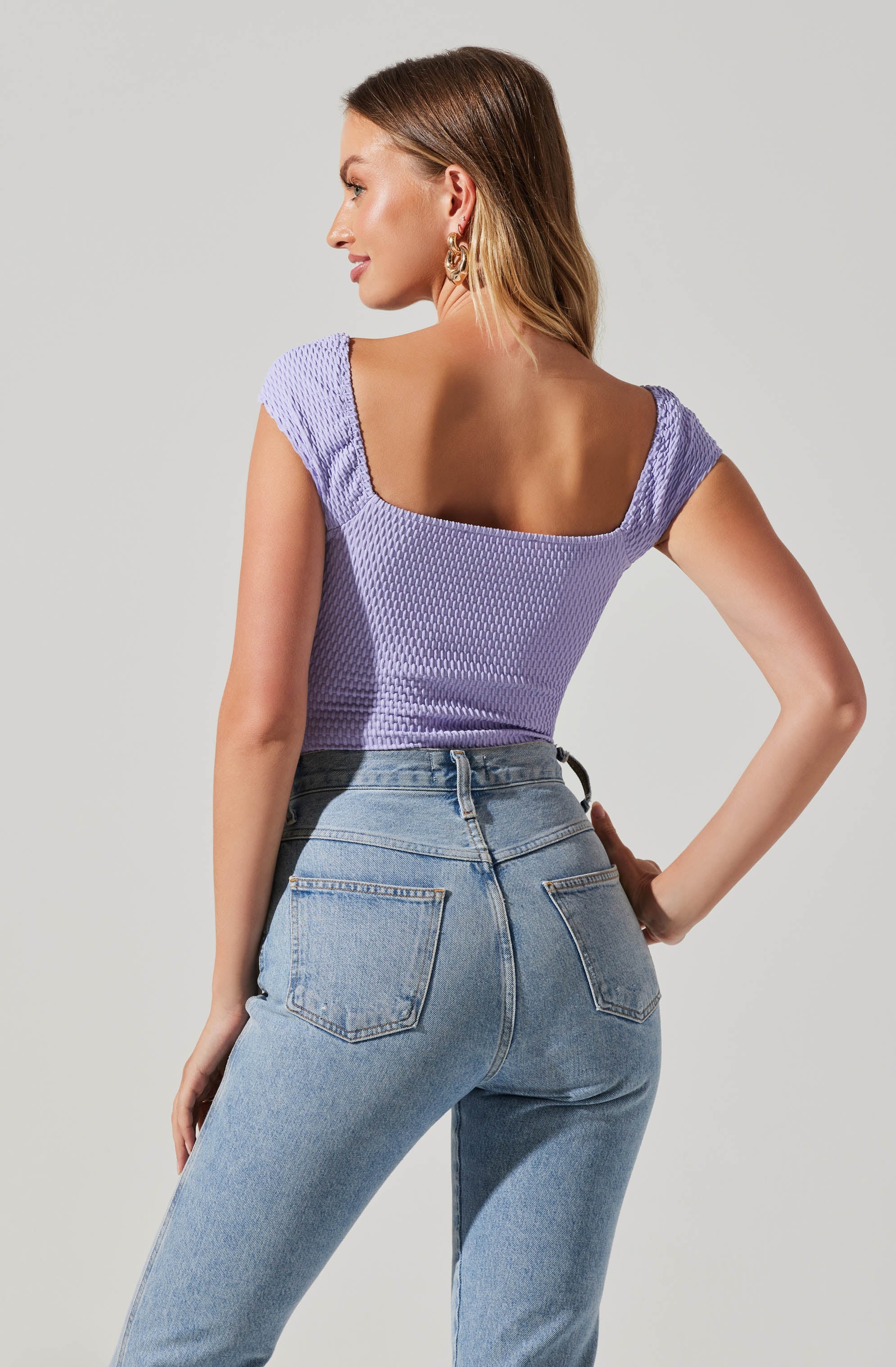 A woman is wearing a lavender, textured, short-sleeve top with a wide square neckline and fitted appearance. She is paired it with high-waisted, light-wash jeans that have two back pockets.