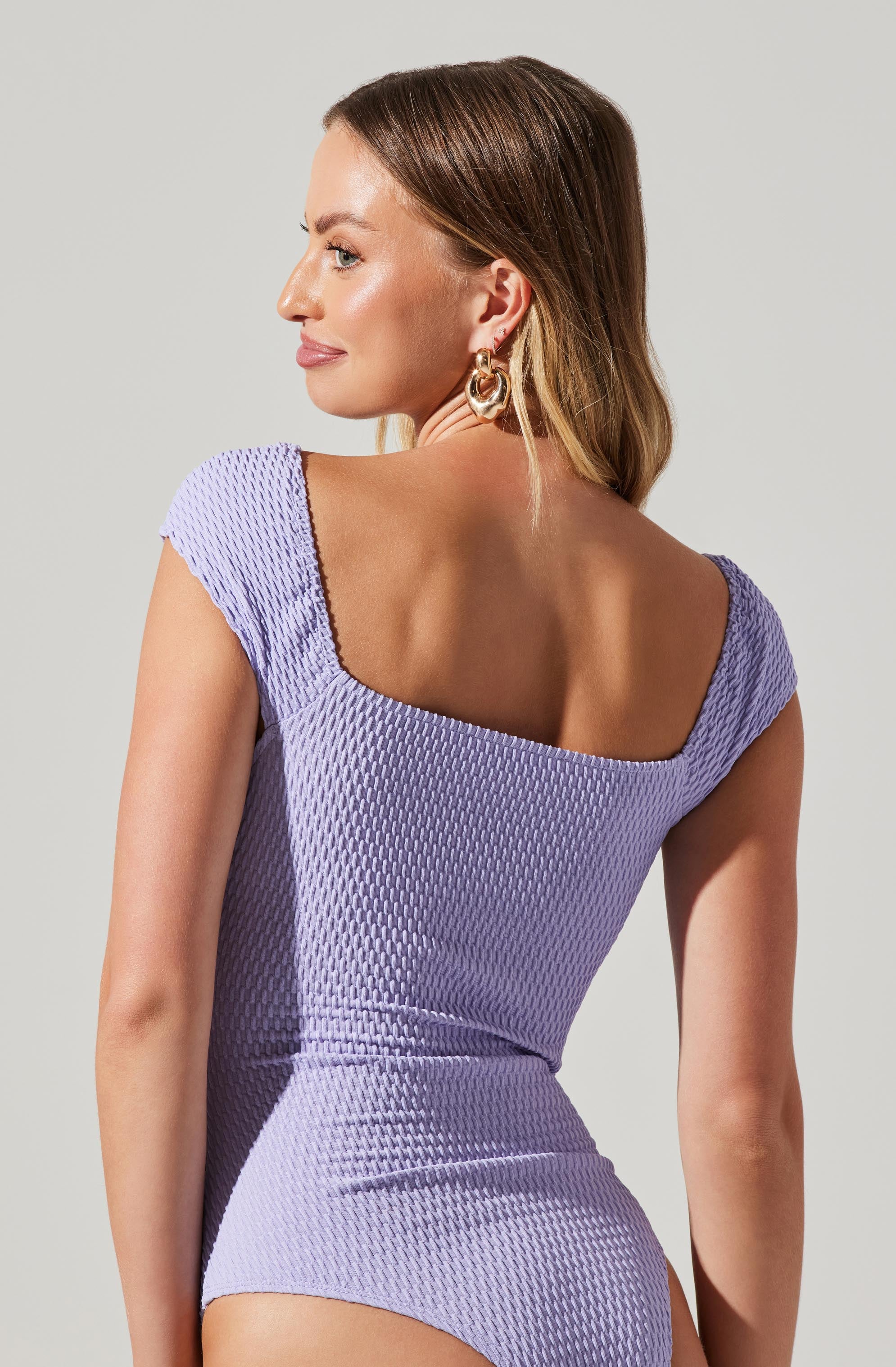 A woman is wearing a lavender-colored textured dress with cap sleeves and a square neckline. The fabric has a small, repeating pattern, giving it a slightly raised, textured appearance. She is accessorized with large, gold hoop earrings.