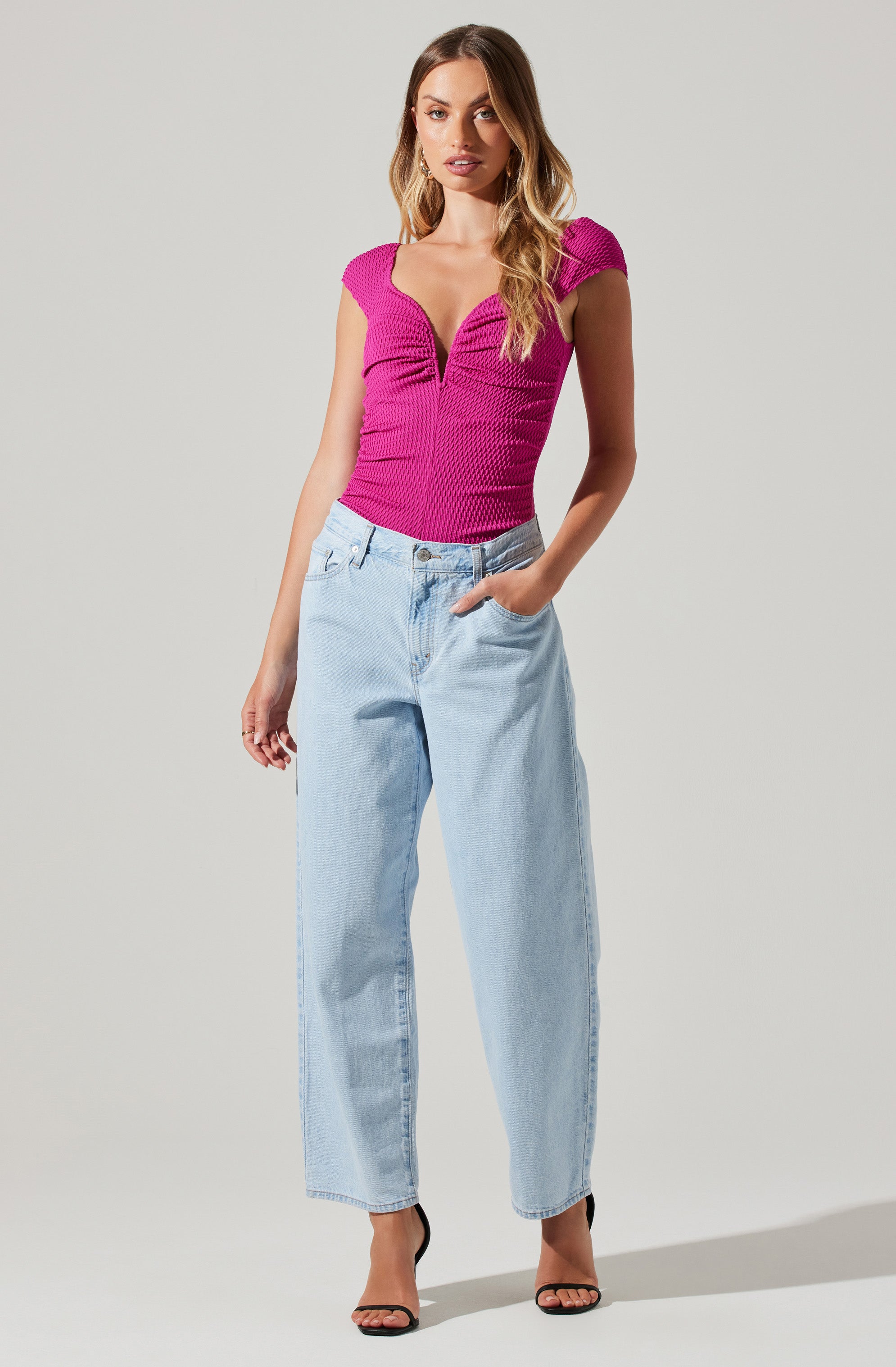 A woman is wearing a fitted, short-sleeved, textured magenta top with a deep V-neckline. She pairs it with high-waisted, light-wash, loose-fitting jeans and black high-heeled sandals.