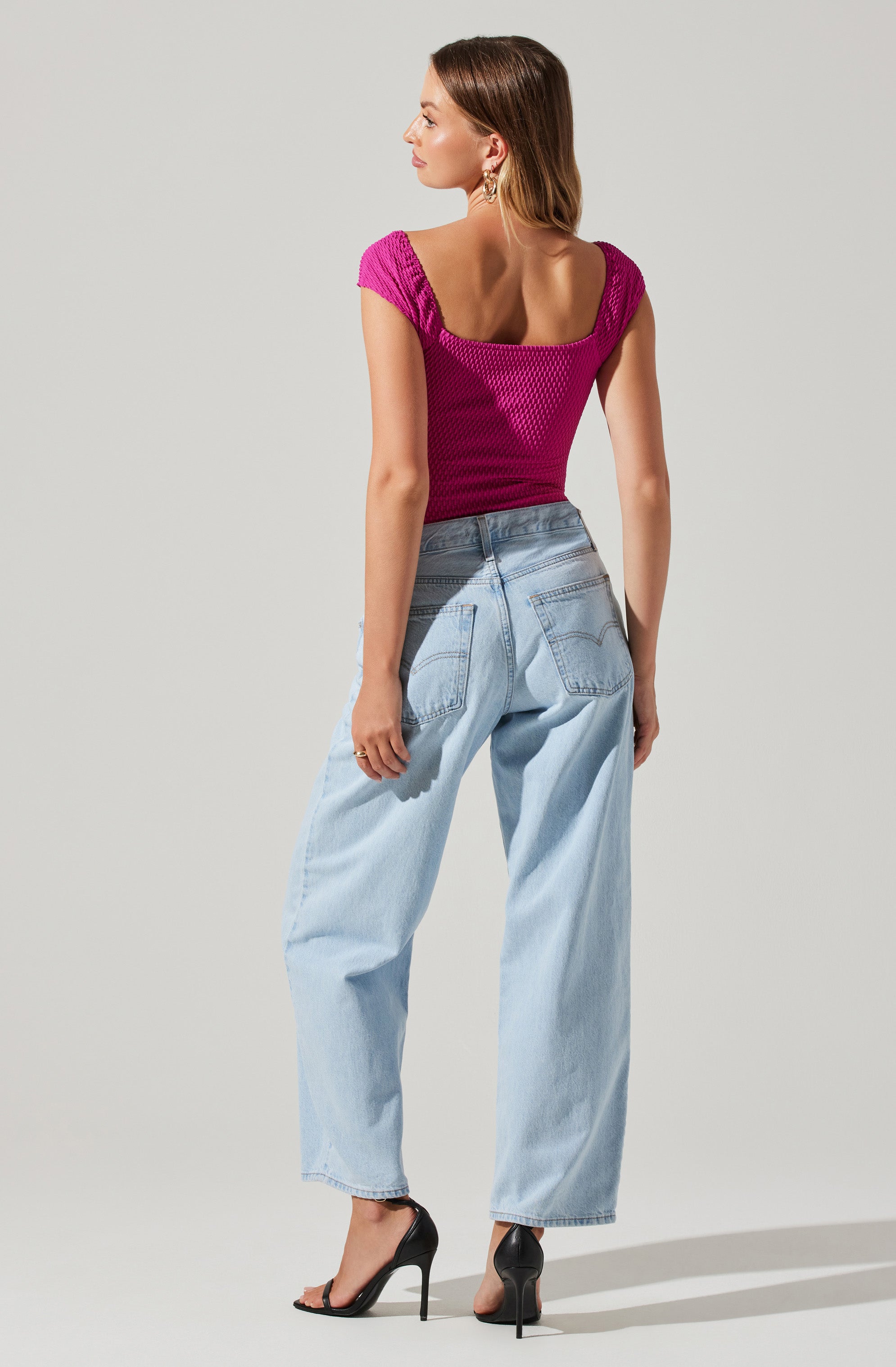 A woman is wearing a fitted, short-sleeved, textured magenta top with a scoop neckline. She pairs it with high-waisted, light-wash, wide-leg jeans and black high-heeled sandals.