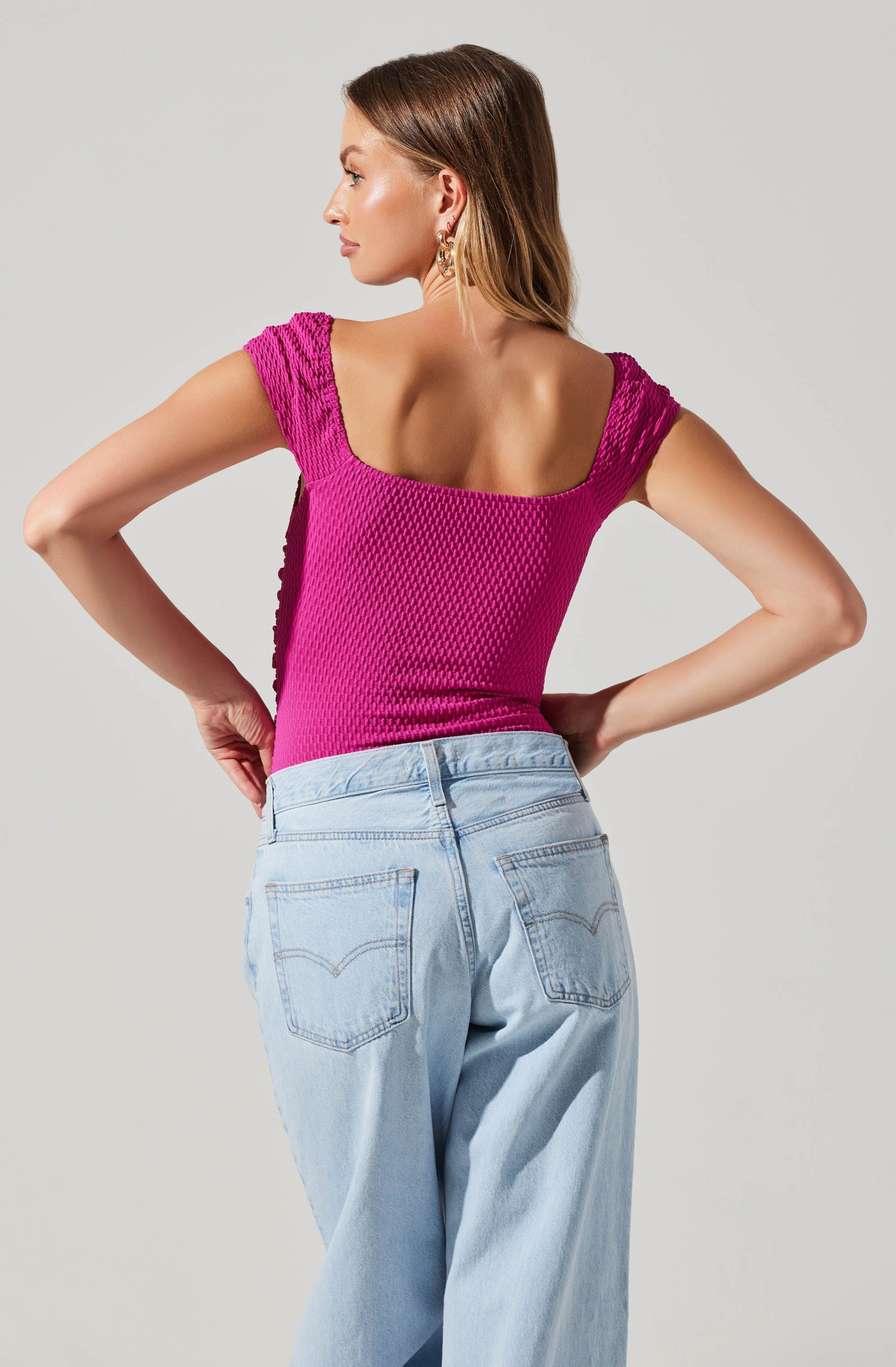 A woman is wearing a bright pink, textured, short-sleeved top with a square neckline and shoulder detailing. She pairs it with light blue, high-waisted jeans featuring visible stitching on the back pockets.