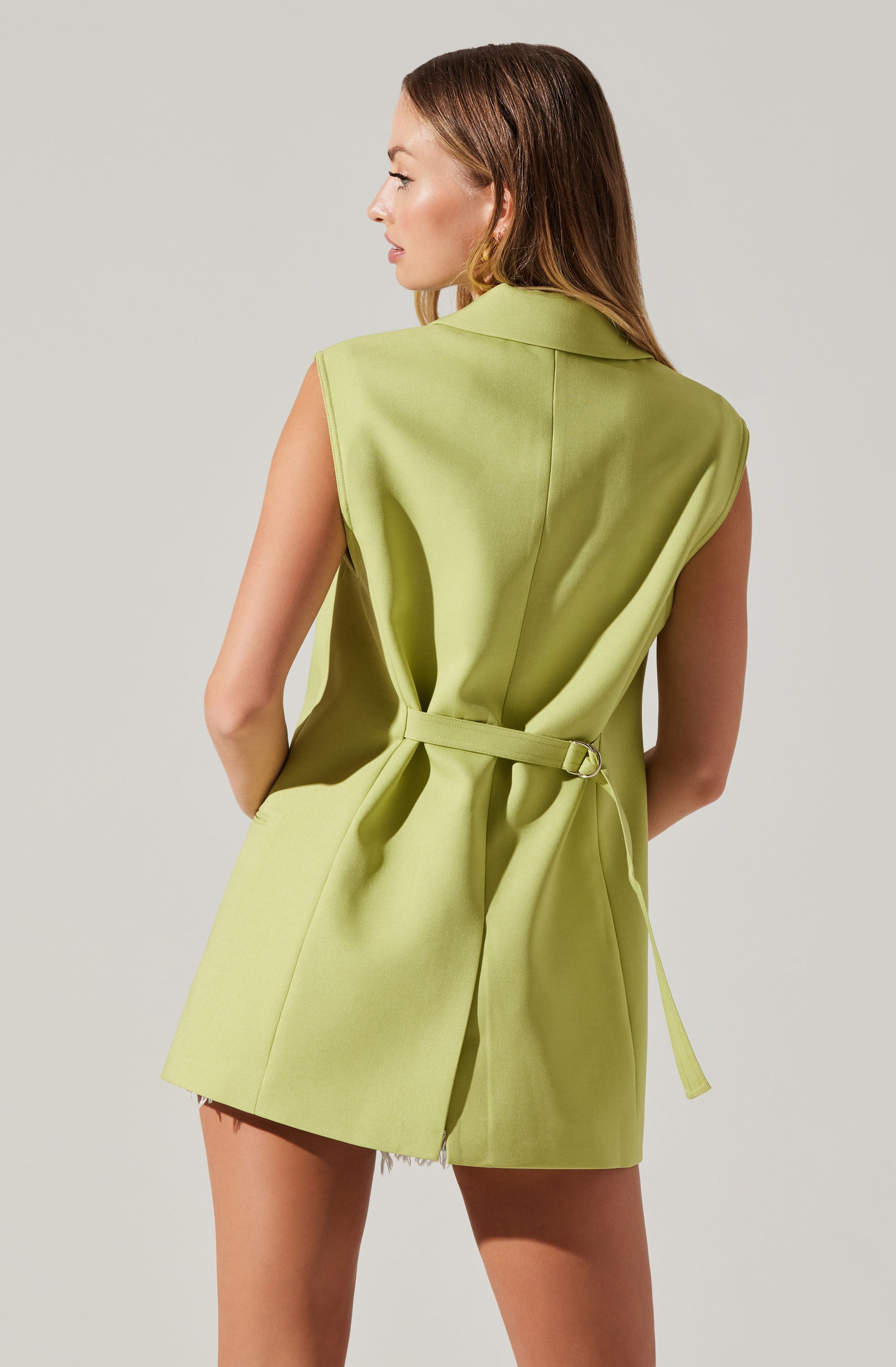 A woman is wearing a sleeveless, lime green dress with a collar and a matching belt at the waist. The dress has a loose fit with some gathered fabric at the waist, creating a draped effect. The hemline falls above the knees.