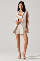 The woman is wearing a sleeveless, beige blazer-style vest with wide shoulder lapels and buttons down the front. She has paired it with matching high-waisted shorts that have a pleated front and a belt. She is also wearing a white cropped top underneath the vest. Her footwear consists of elegant, white high-heeled sandals with ankle straps.