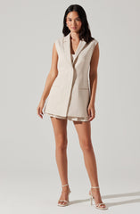 A woman is wearing a sleeveless, beige tailored vest with a notched lapel collar and front pockets. She pairs it with matching beige open-toe high-heeled sandals.