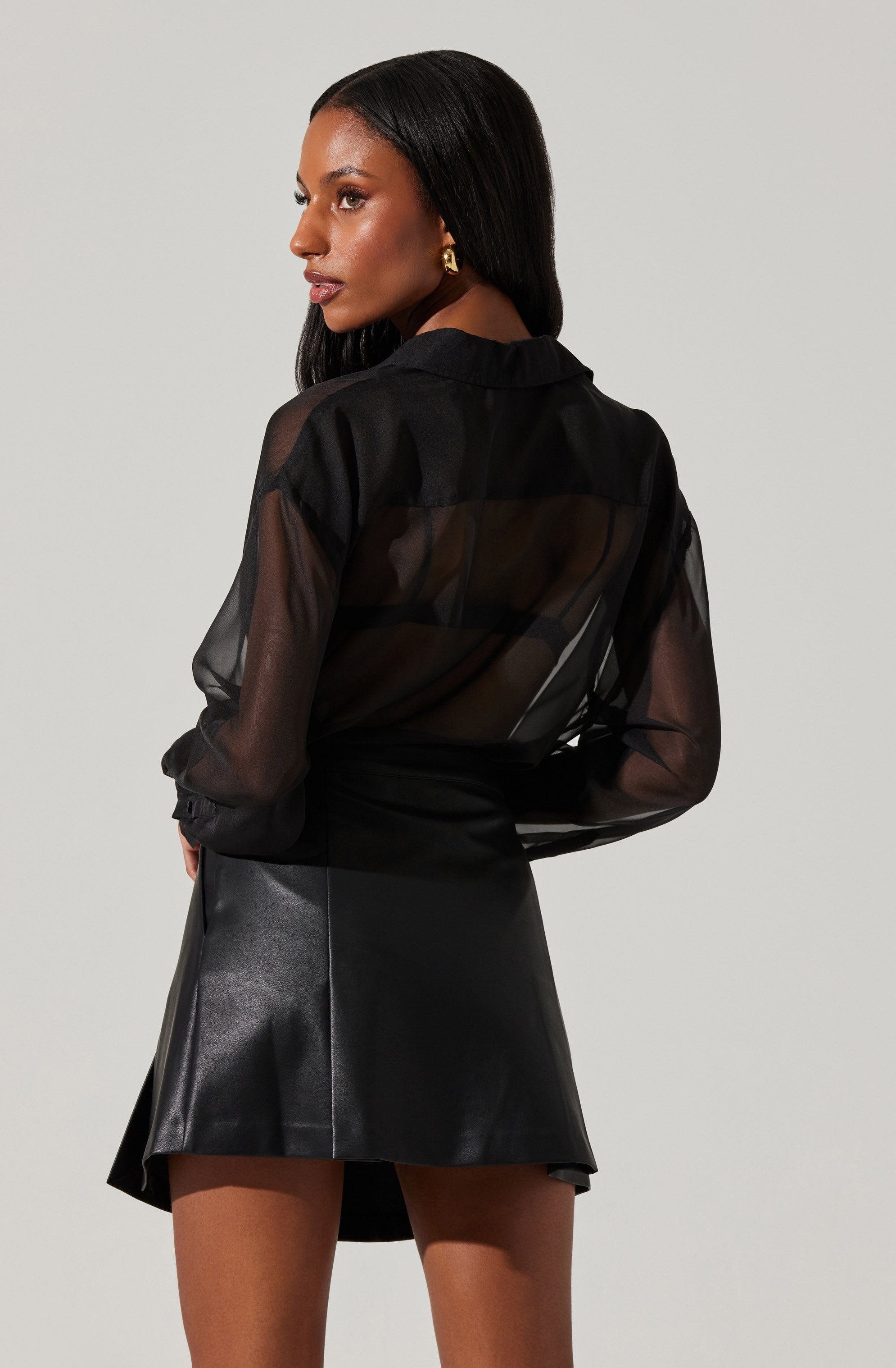 A woman is wearing a sheer black long-sleeve blouse with a collar. The blouse is see-through, revealing a black top underneath. She pairs it with a high-waisted black leather skirt that falls above the knees.