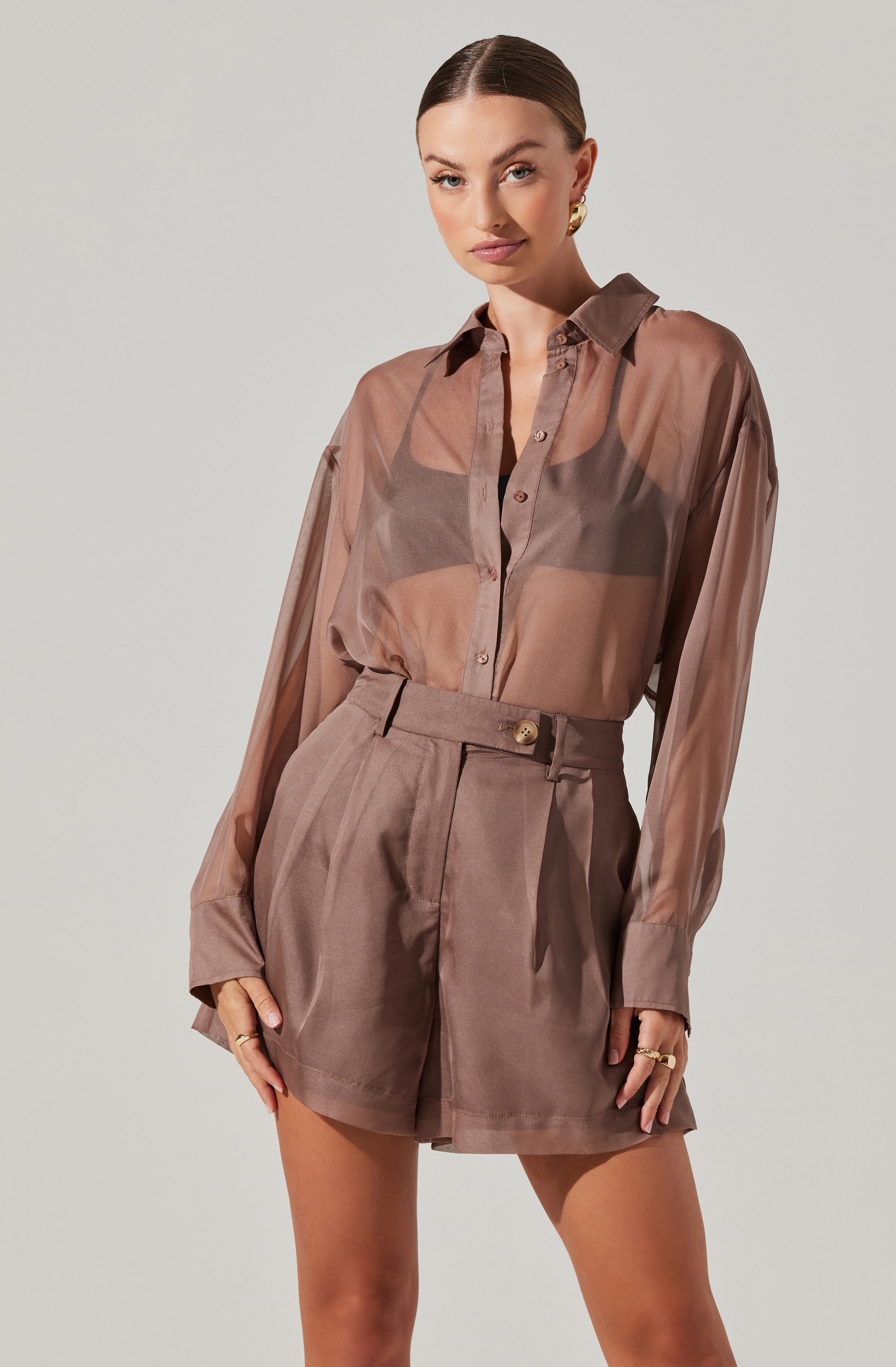 A woman is wearing a sheer, long-sleeved button-up shirt in a soft taupe color, paired with high-waisted shorts in a matching taupe shade. The shirt appears lightweight and slightly translucent, with a classic collar and buttons down the front. The shorts have pleats, a belt with a gold button, and reach mid-thigh.