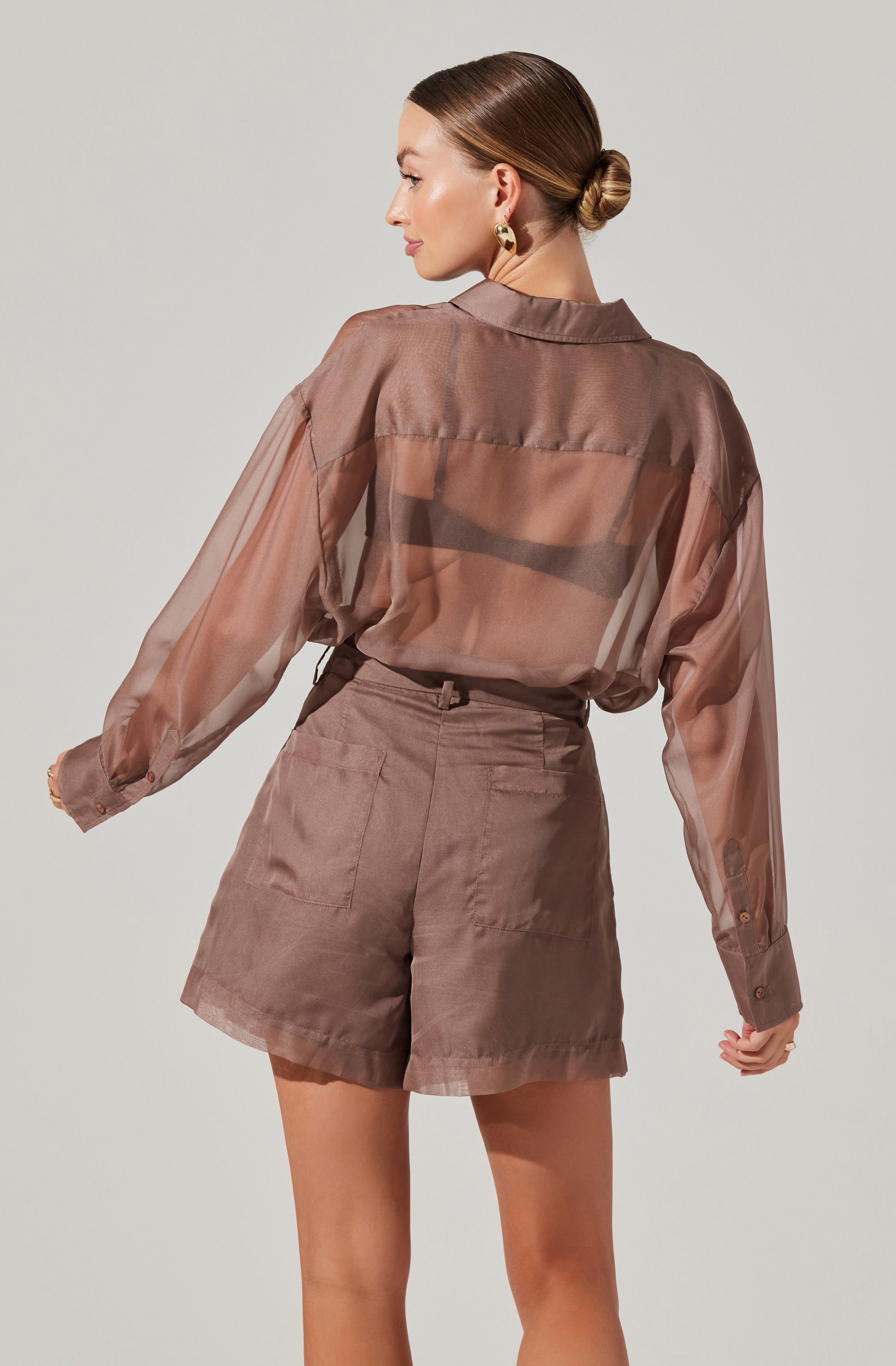 A woman is wearing a sheer, brown long-sleeve button-up blouse with a classic collar. The blouse is translucent, revealing a darker undergarment underneath. She pairs it with high-waisted, loose-fitting shorts in a matching brown shade, featuring large patch pockets on the back. The outfit has a neutral and sophisticated tone.
