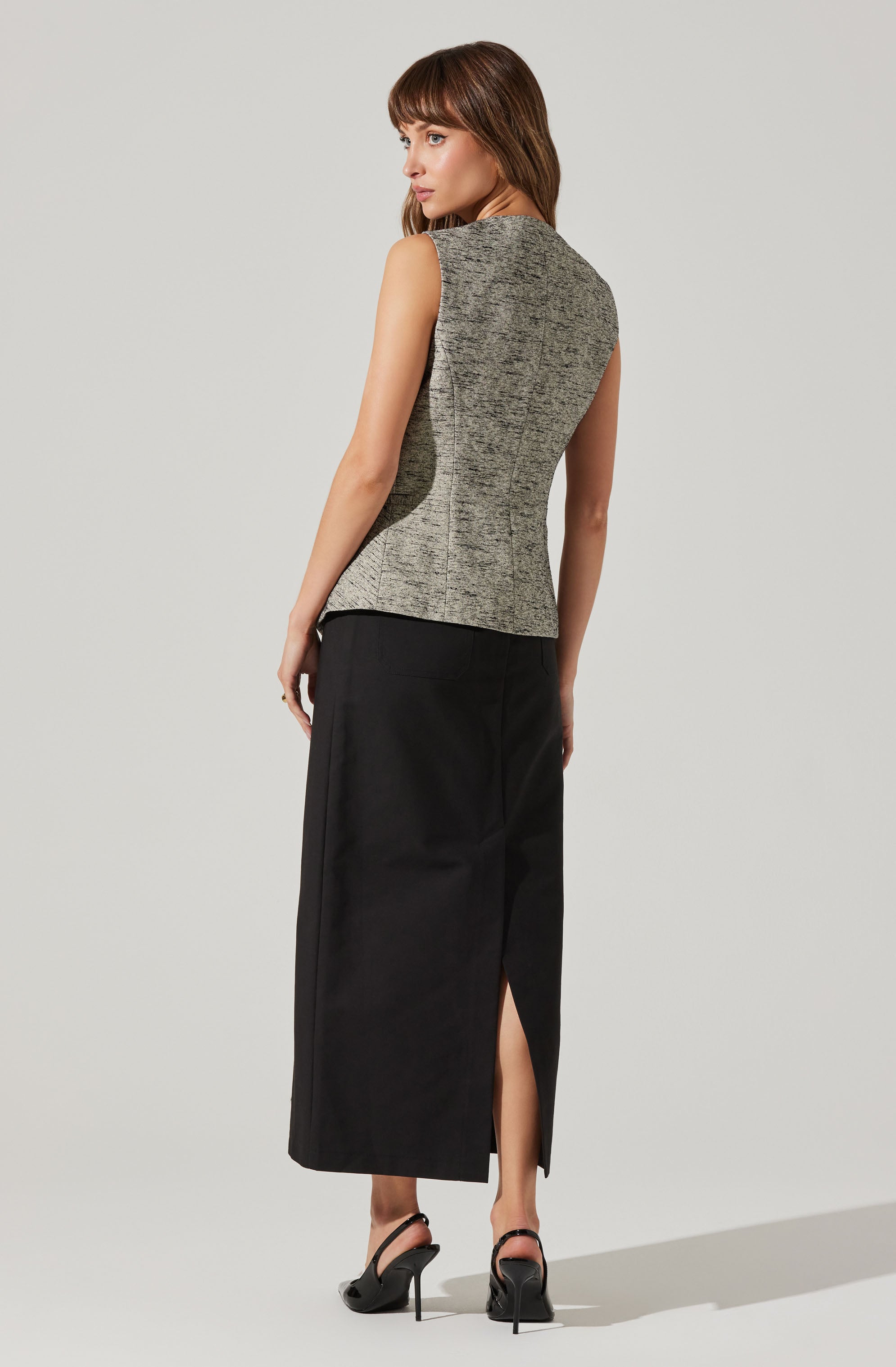 A woman is wearing a sleeveless, fitted, gray textured top with a round neckline. She pairs it with a high-waisted, black, ankle-length skirt that features a front slit. She completes her outfit with black high-heeled sandals.