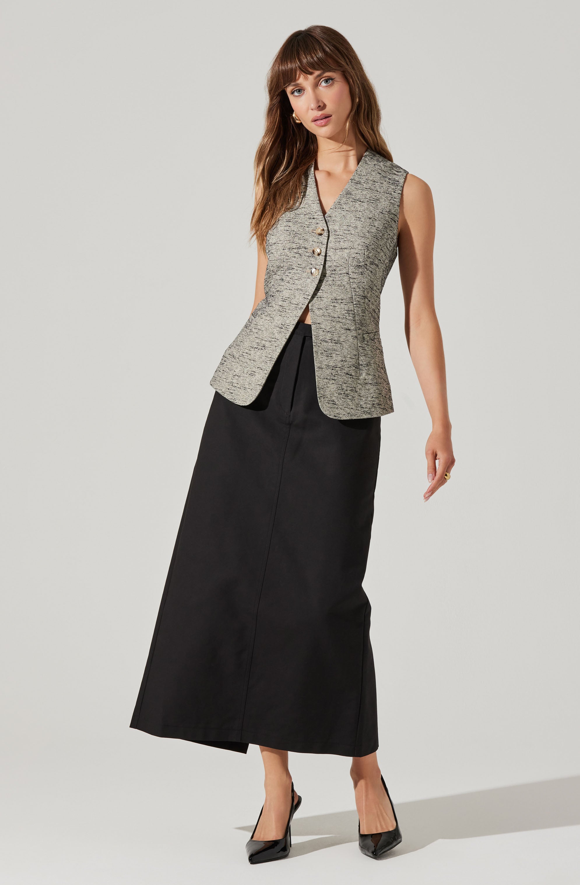 A woman is wearing a sleeveless, v-neck, gray textured vest with metallic buttons down the front. She is also wearing a long, high-waisted black skirt that reaches her ankles, paired with black pointed-toe high heels.