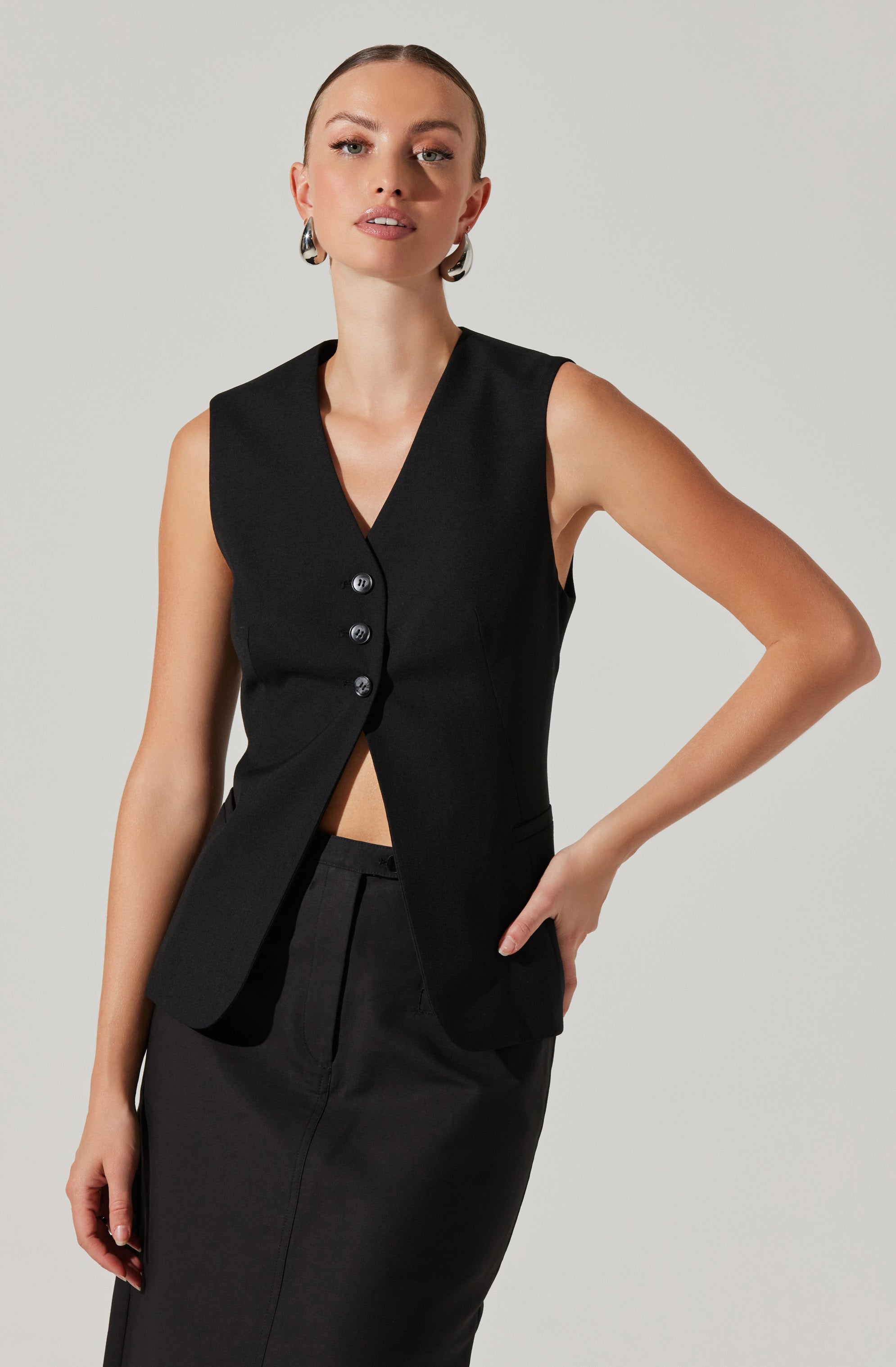A woman is wearing a sleeveless black vest with a deep V-neck and four buttons down the front. The vest is fitted and tailored, with a slightly flared hem. She pairs it with a matching black skirt that has a high waist and a sleek, straight silhouette. She also has large, statement earrings.