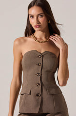 A woman is wearing a strapless, fitted blazer with a houndstooth pattern in shades of brown and black. The blazer features large decorative buttons down the front and two flap pockets at the waist. She accessorizes with a chunky gold chain necklace, gold earrings, and a gold ring.