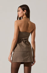 A woman is wearing a strapless, fitted dress with a subtle checkered pattern in neutral tones. The dress is cinched at the waist with a small belt. She has accessorized with a chunky gold chain necklace and large gold earrings.