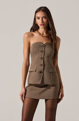 A woman is wearing a strapless, tailored, brown checkered mini dress with large front buttons and two flap pockets at the hips. She is accessorized with a chunky gold chain necklace, gold hoop earrings, and a gold ring on her right hand. She is also wearing sheer black tights.